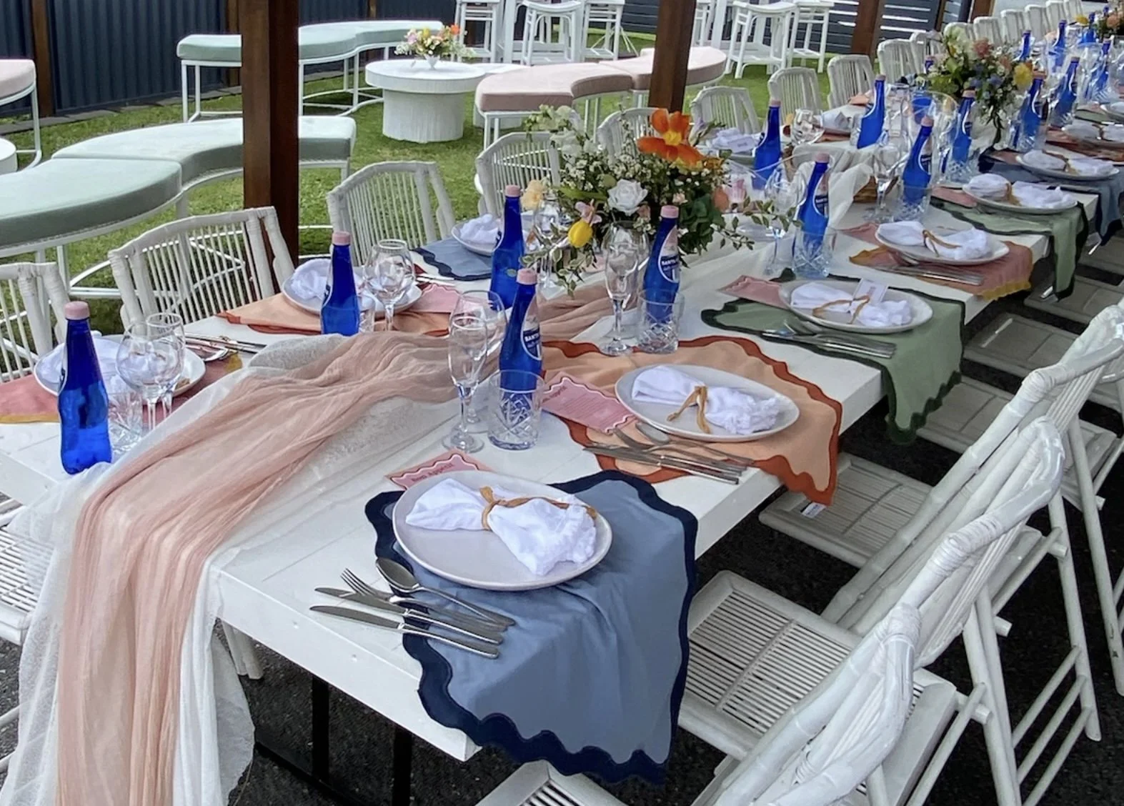 A long table set for an outdoor event with floral centerpieces, colorful table runners, white plates, napkins, glassware, and blue bottles of water, under a pergola.