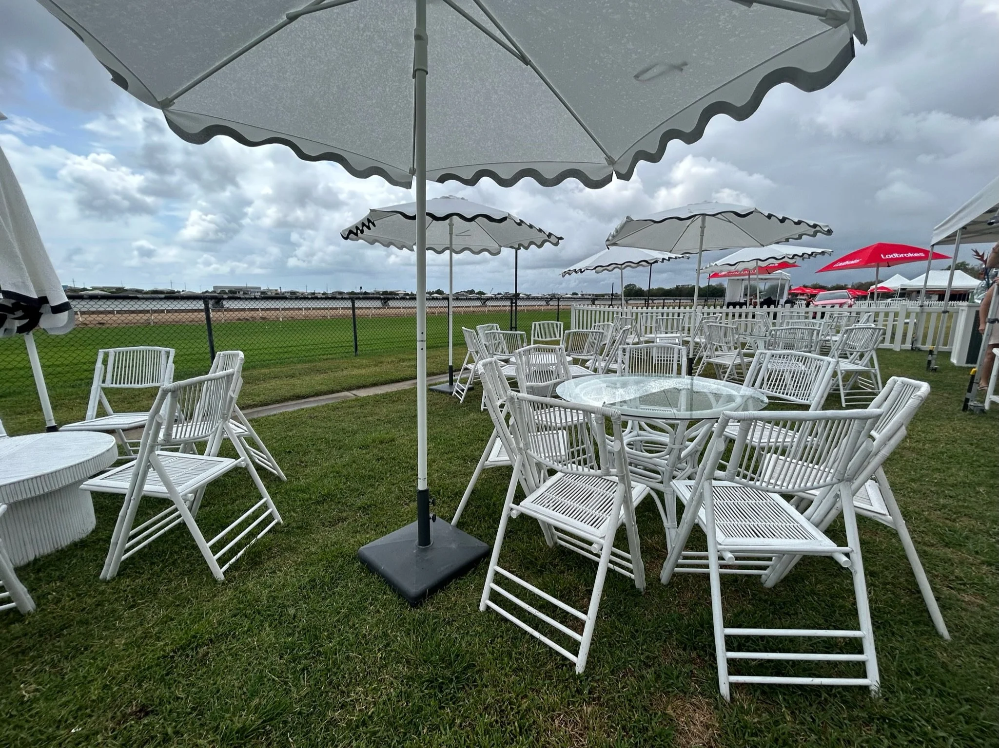 Empty outdoor seating area with white chairs and glass tables, shaded by large white umbrellas on a grassy field, overcast sky, and a race track in the background.