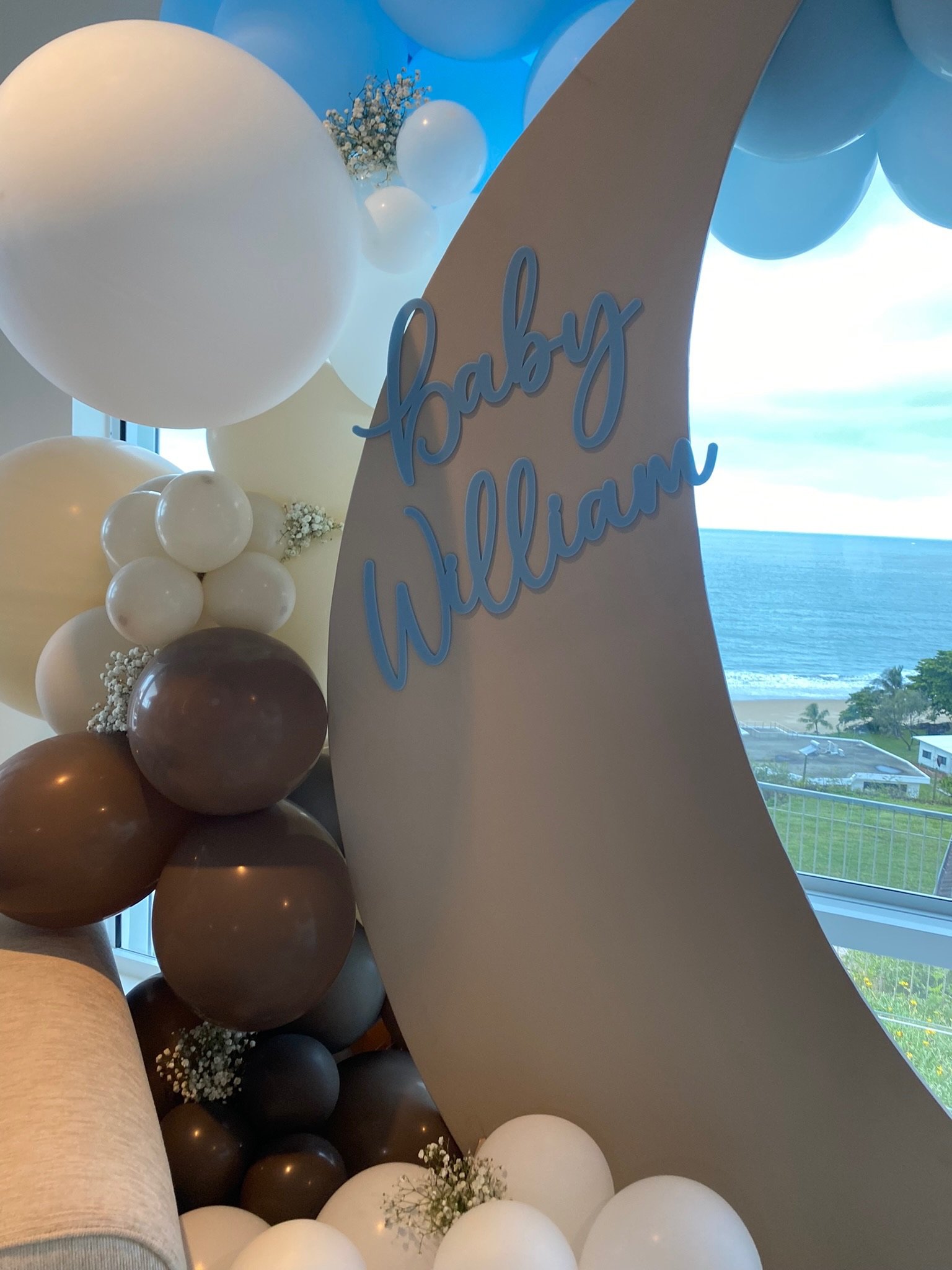 Decorative balloon display with white, cream, and brown balloons, baby's breath flowers, and a large gray crescent-shaped panel with blue cursive text that reads 'Baby William'. There is a window showing an ocean view outside.