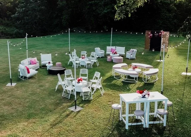 Outdoor party setup with white tables, chairs, and sofas on a grassy lawn, decorated with pink accents and string lights, surrounded by trees.