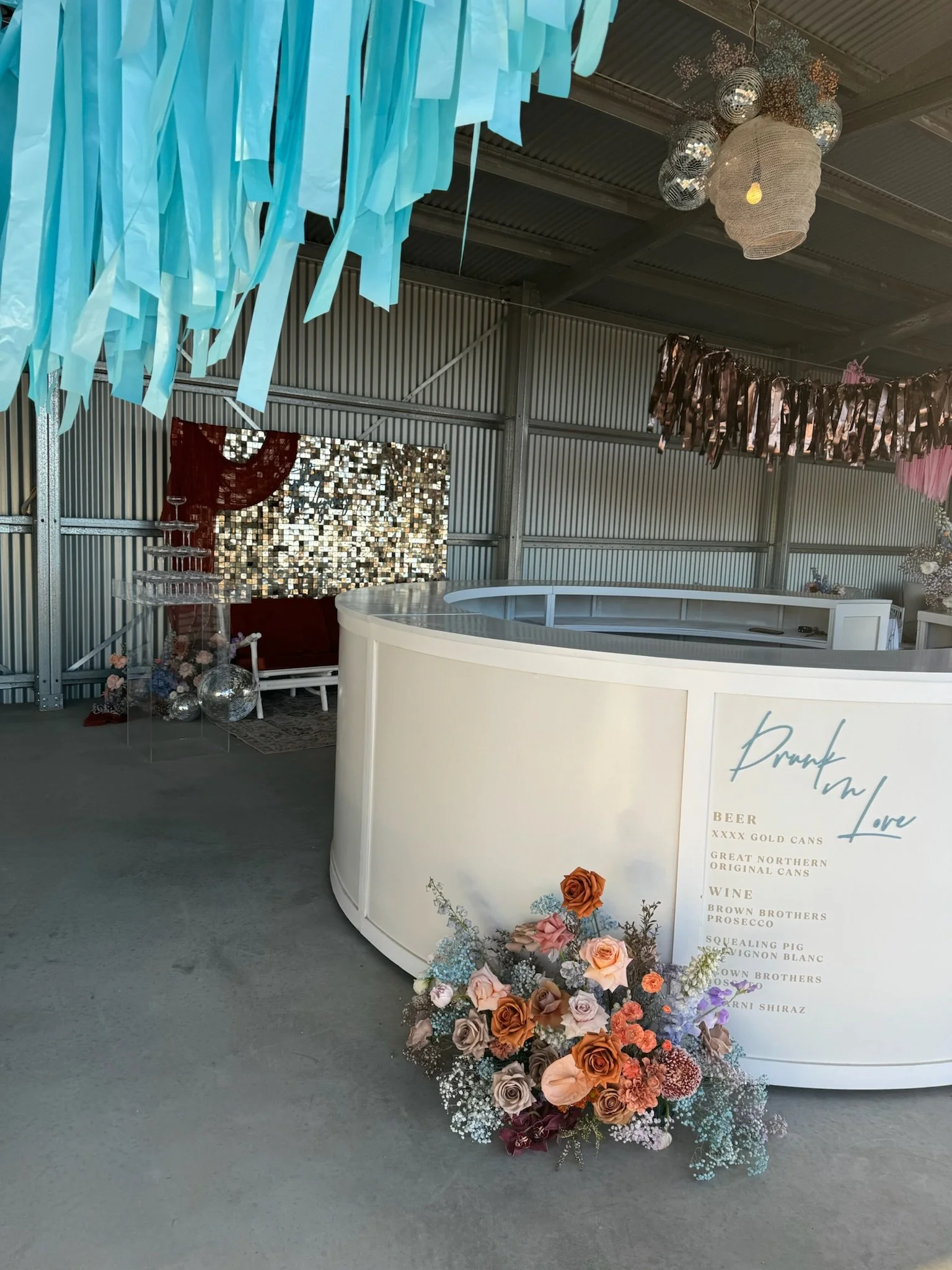 Decorative event space with a white bar, floral arrangements, hanging blue streamers, mirrored disco balls, and a metallic backdrop.