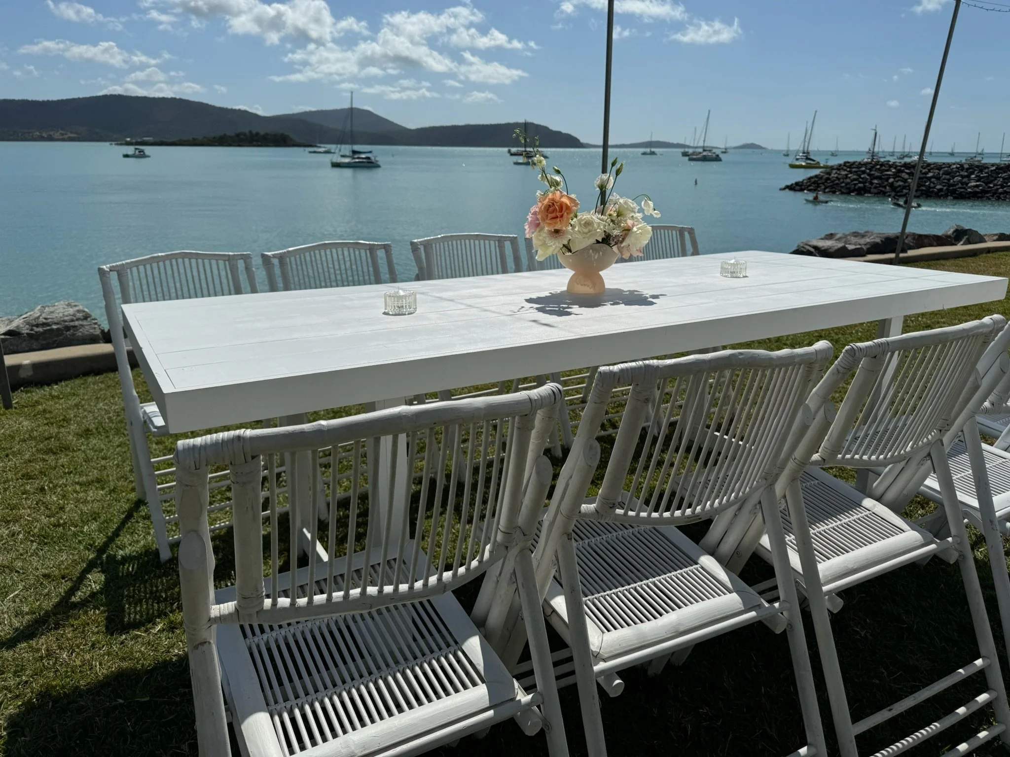 White dining table with white chairs on grass by the water, decorated with a flower arrangement and small glass candle holders, overlooking boats and mountains in the distance.