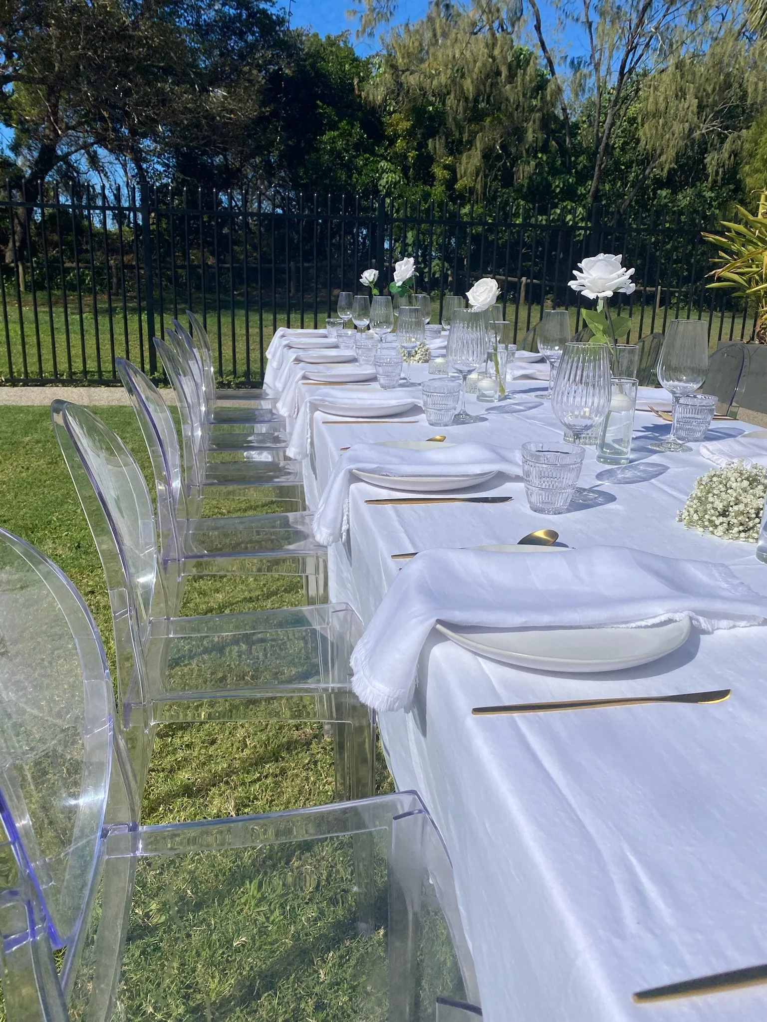 Outdoor dining table set with white tablecloth, clear chairs, white flowers, and glassware on a sunny day