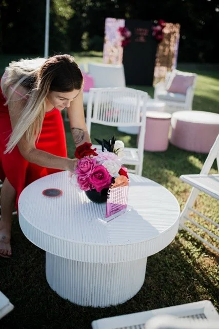 A woman in a red dress arranging flowers in a bouquet on a white outdoor table at a garden event.