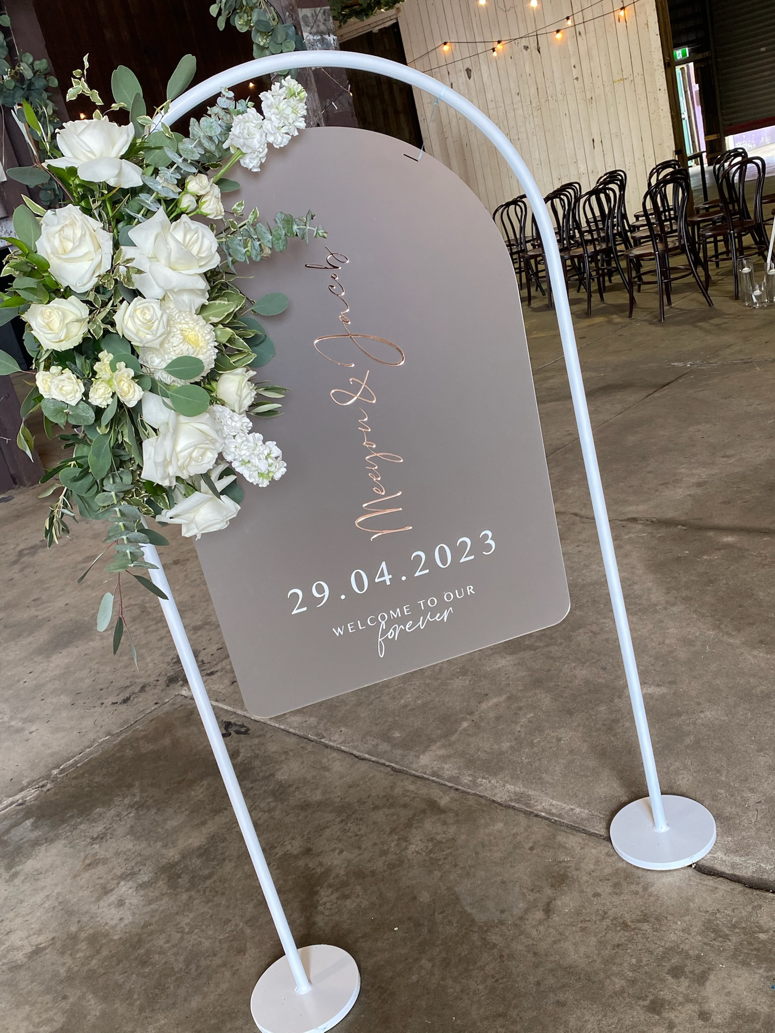 Wedding welcome sign with white and green floral arrangements on a white stand, displaying the date 29.04.2023 and the message 'Welcome to our forever'