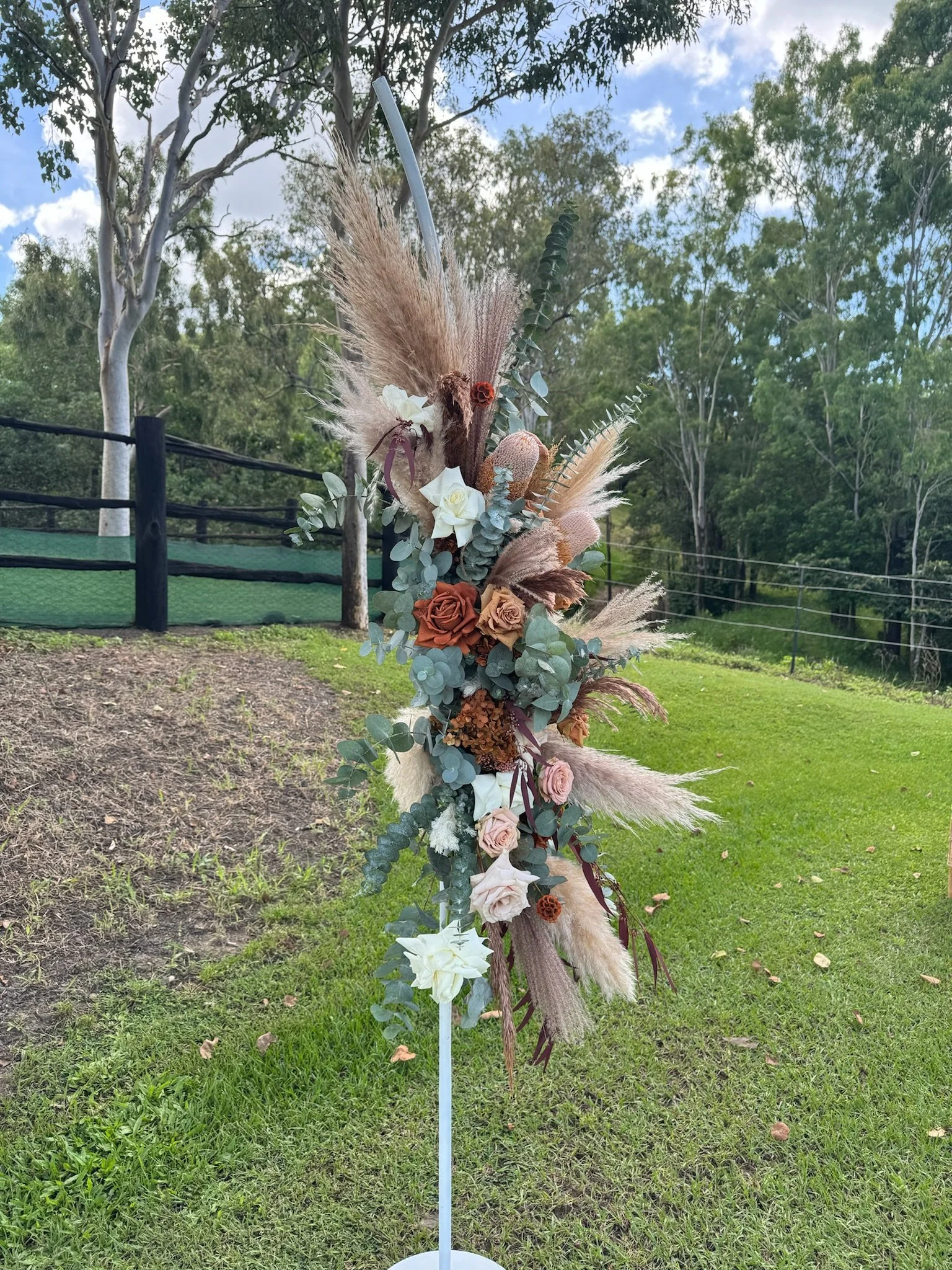 A freestanding floral arrangement with a mix of dried and fresh flowers in neutral tones, including white roses, pink roses, eucalyptus, pampas grass, and other dried grasses, set outdoors on grass with trees and a fence in the background.