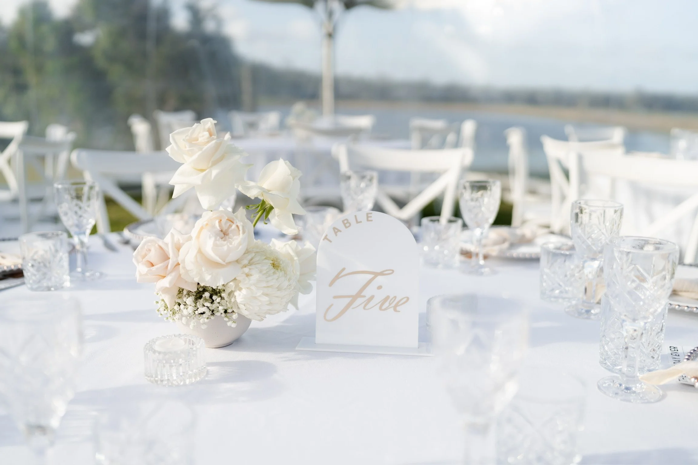 Elegant outdoor wedding reception table decorated with white flowers, glassware, and a table number sign reading 'Five', with a lakeside view in the background.