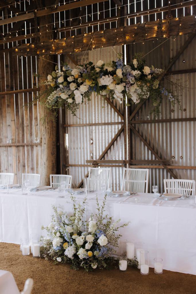 Decorated rustic barn wedding reception with a long table, white tablecloth, white chairs, floral arrangements, candles, and string lights.