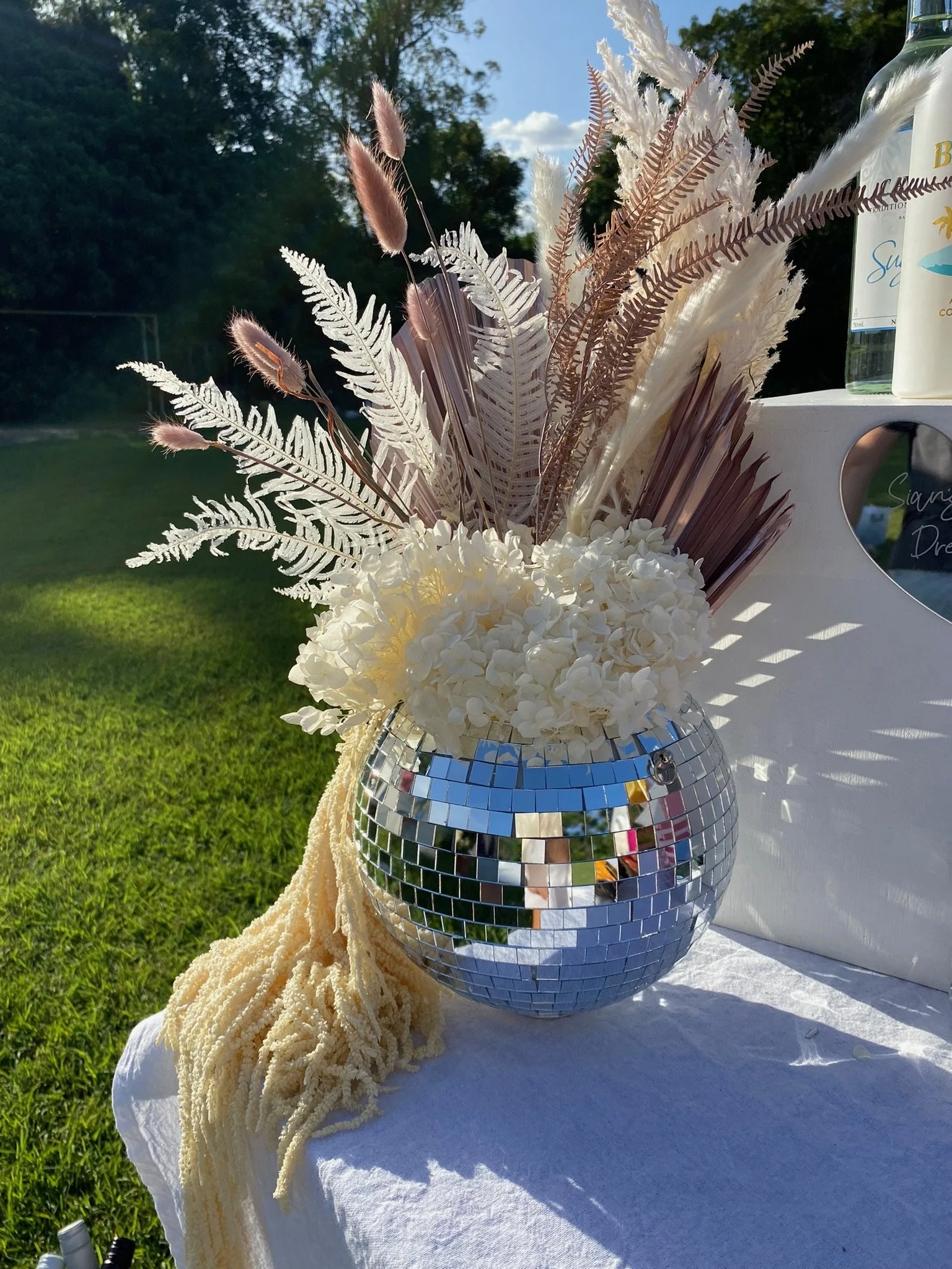 Decorative floral arrangement in a mirror ball vase with white hydrangeas, dried ferns, pampas grass, and other dried flowers on a table outdoors with grass and trees in the background.