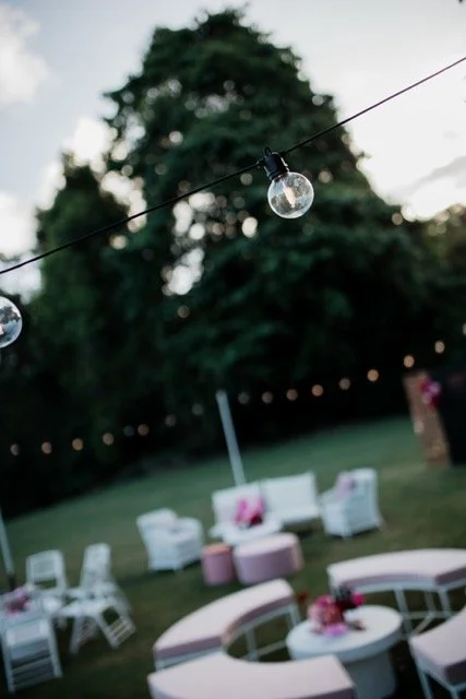Outdoor party setup with string lights, empty chairs and tables, and a large tree in the background.