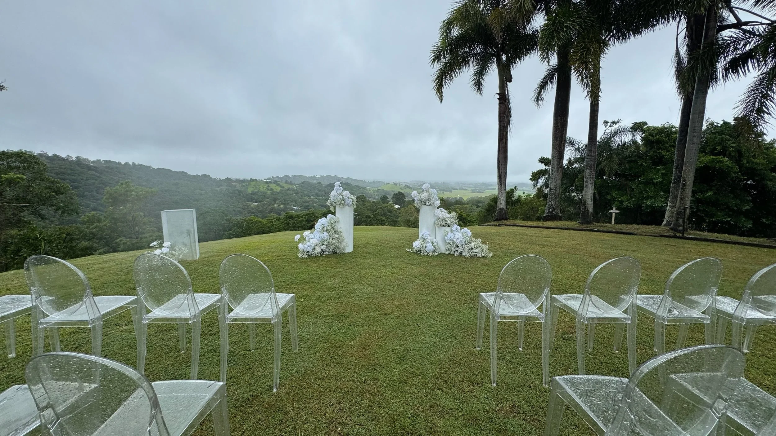 Outdoor wedding setup with transparent chairs on a grassy hill, white floral arrangements on high pedestals, and tall palm trees, with a scenic view of rolling hills under a cloudy sky.