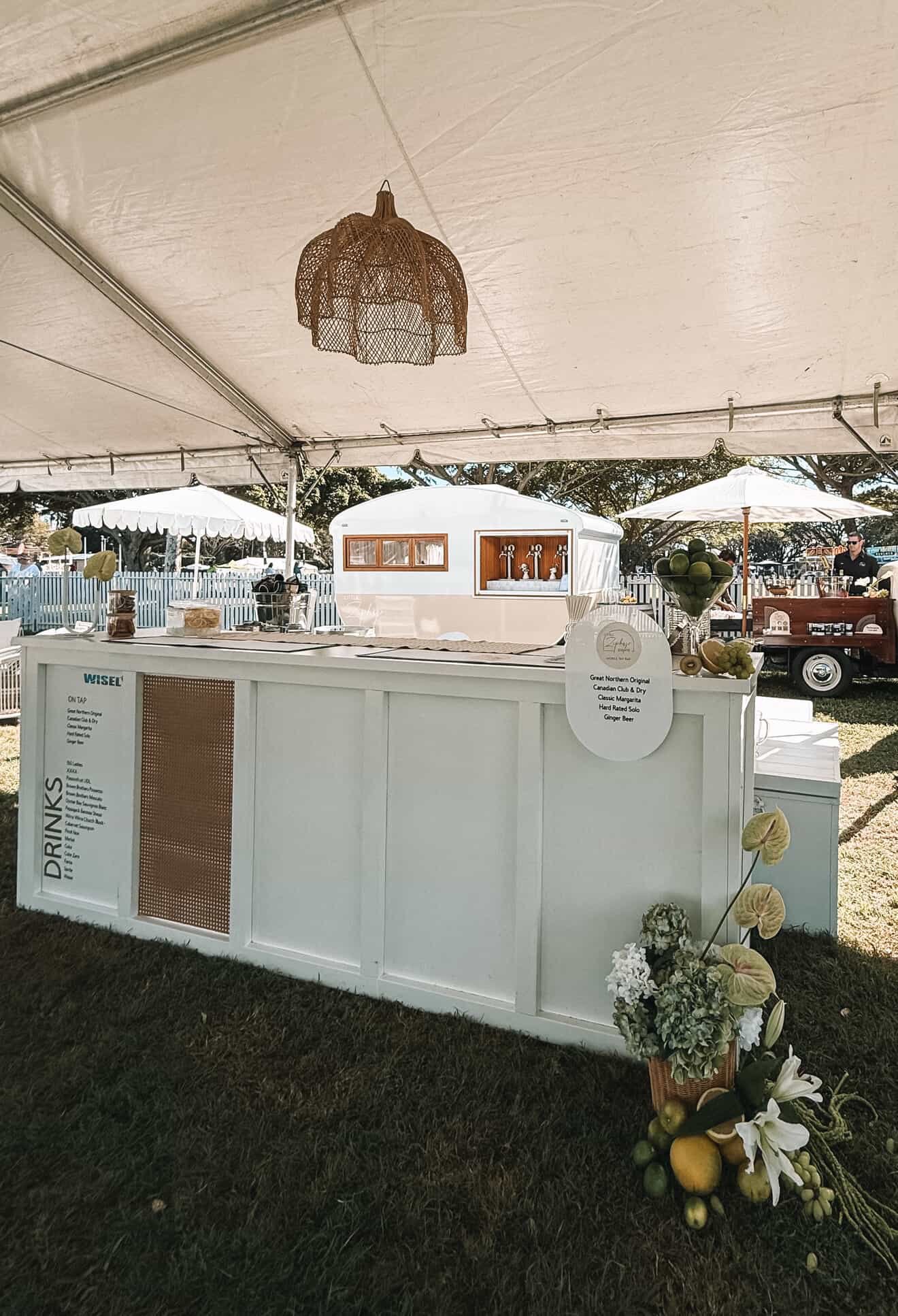 A white outdoor beverage stand under a tent at a fair or event, with fruit decorations and a menu sign, and a small vintage trailer in the background.