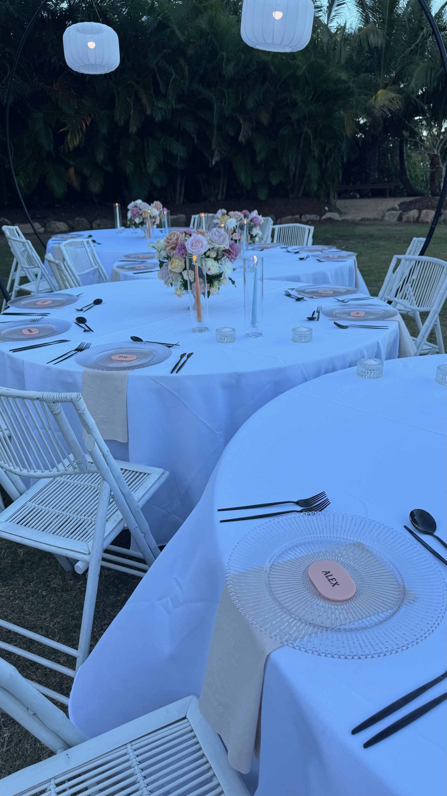 Outdoor dining setup with white tables, white chairs, floral centerpieces, candles, and place settings with black cutlery and clear glass plates, appearing to be prepared for an event or celebration during the evening.