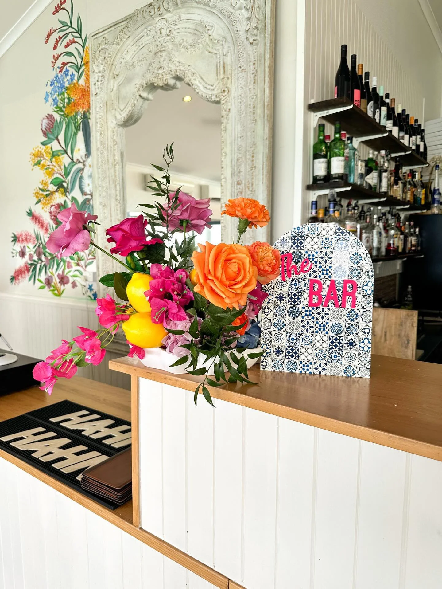Colorful floral arrangement with pink, orange, yellow, and purple flowers on a bar counter with a decorative sign that reads "The Bar." Shelves of liquor bottles are visible in the background, along with a white ornate mirror and a mural of colorful 