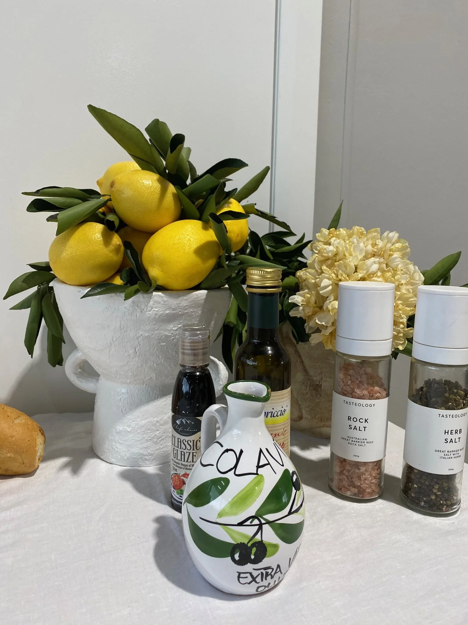 A still life display of yellow lemons with green leaves in a white ceramic vase, surrounded by various bottles of oils and seasonings, a white ceramic pitcher with painted green olives and the words 'Calavr' and 'Exira', with hydrangea flowers in the