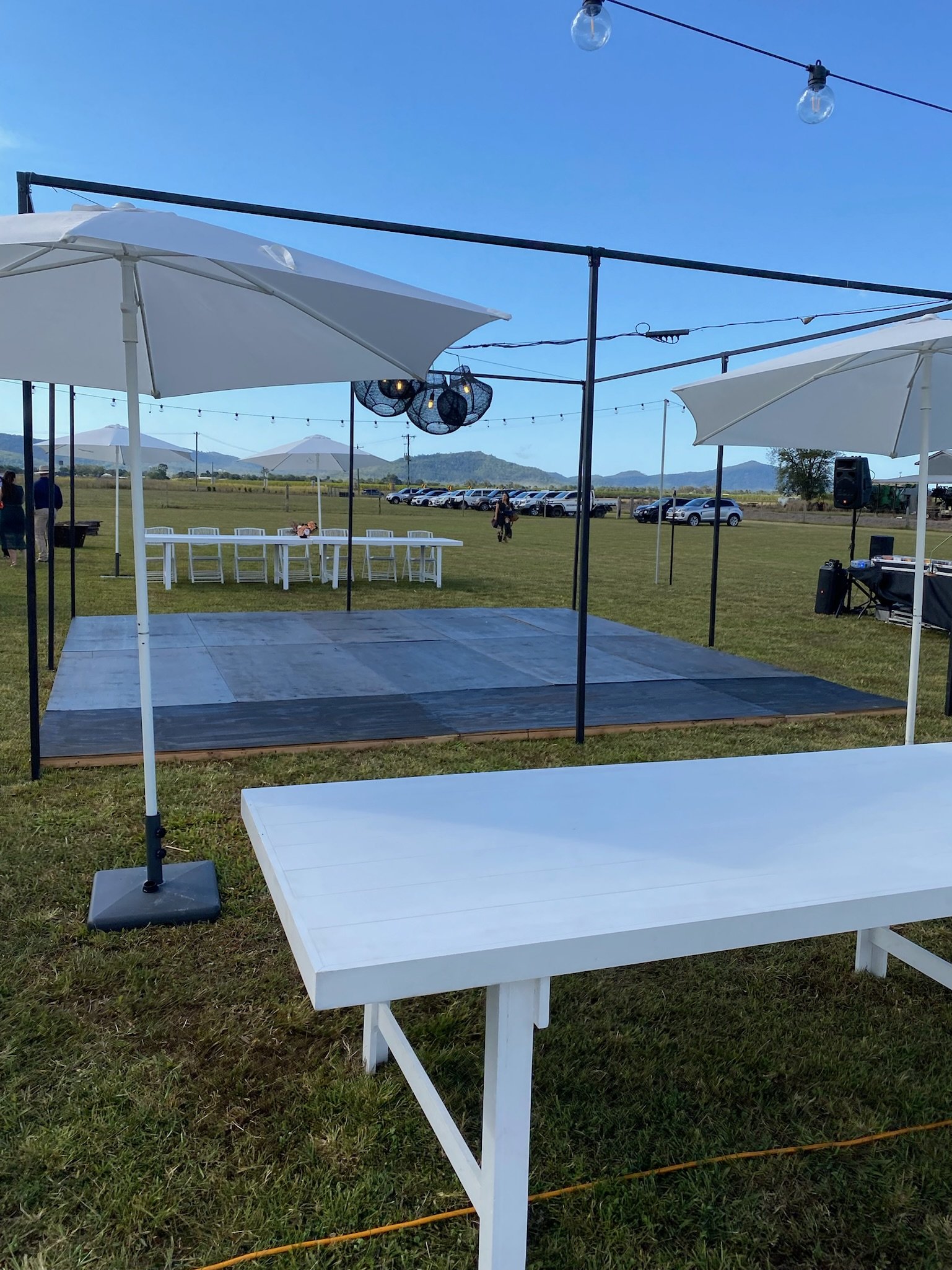Outdoor event space with a small elevated dance floor, white tables, large umbrellas, string lights, and parked cars in the background under a clear blue sky.