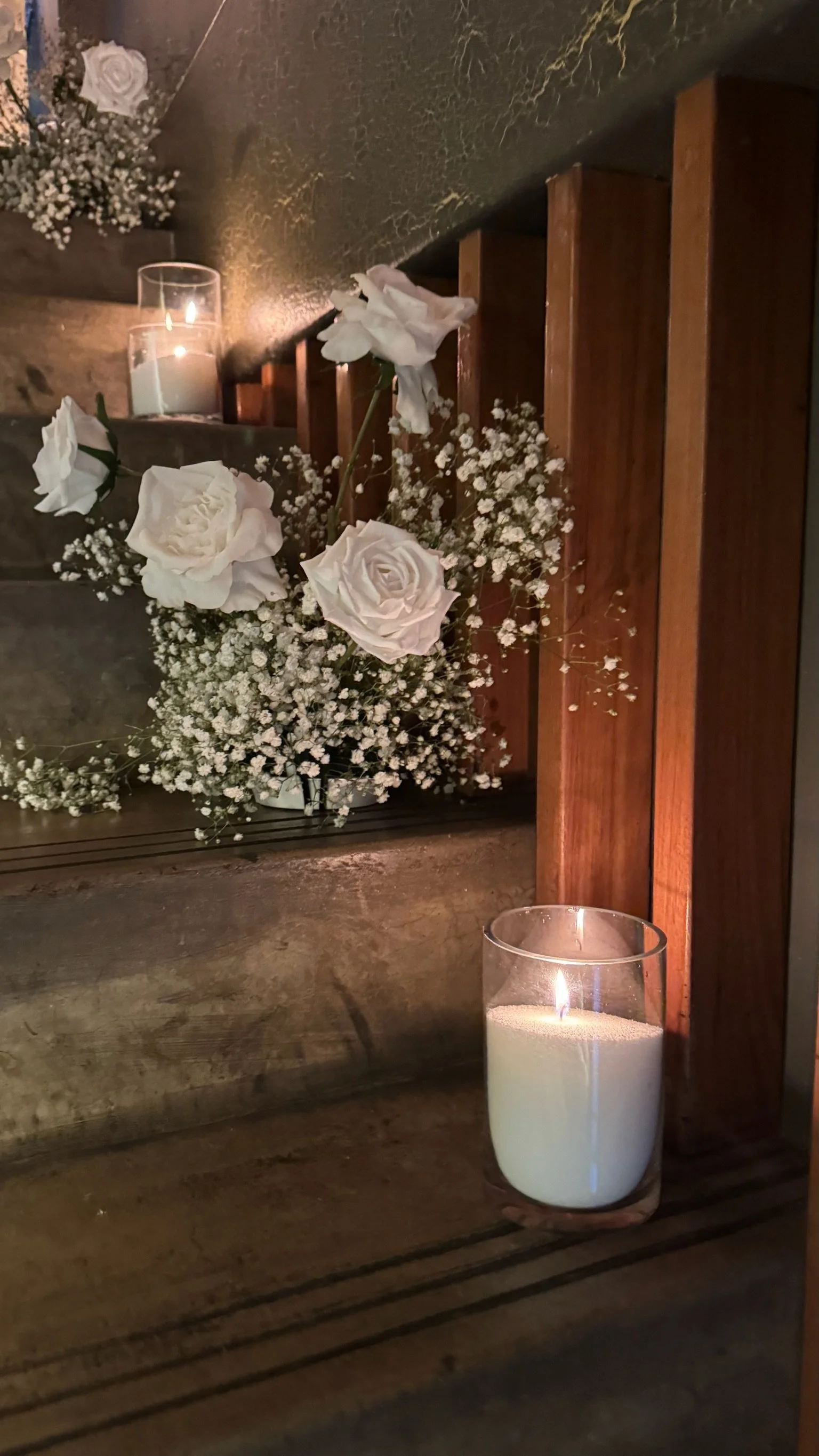 A floral arrangement with white roses and baby's breath on wooden stairs, illuminated by candles.