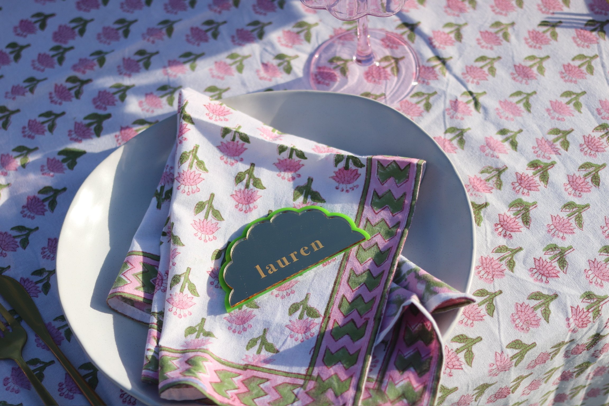 A white plate with a folded napkin that has a pink, green, and white floral pattern, placed on a tablecloth with a matching floral pattern. A name tag with the name "Lauren" is resting on the napkin. There is also a pink wine glass in the background.