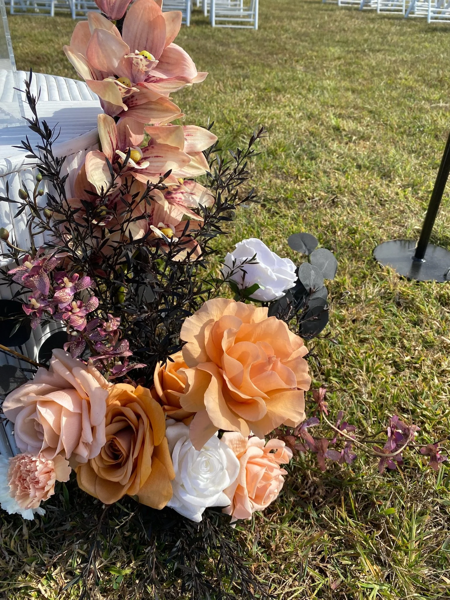 Arrangement of artificial flowers, including roses, orchids, and greenery, placed on grass with chairs in the background at an outdoor event.