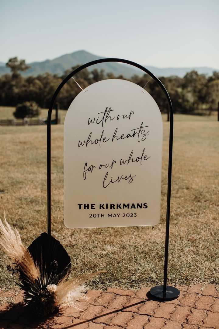 A wedding sign with a white background and black text on a metal stand outdoors in a grassy area with mountains in the distance. The sign reads, "Without our whole hearts for our whole lives." The bottom of the sign has "The Kirkmans 20th May 2023." 