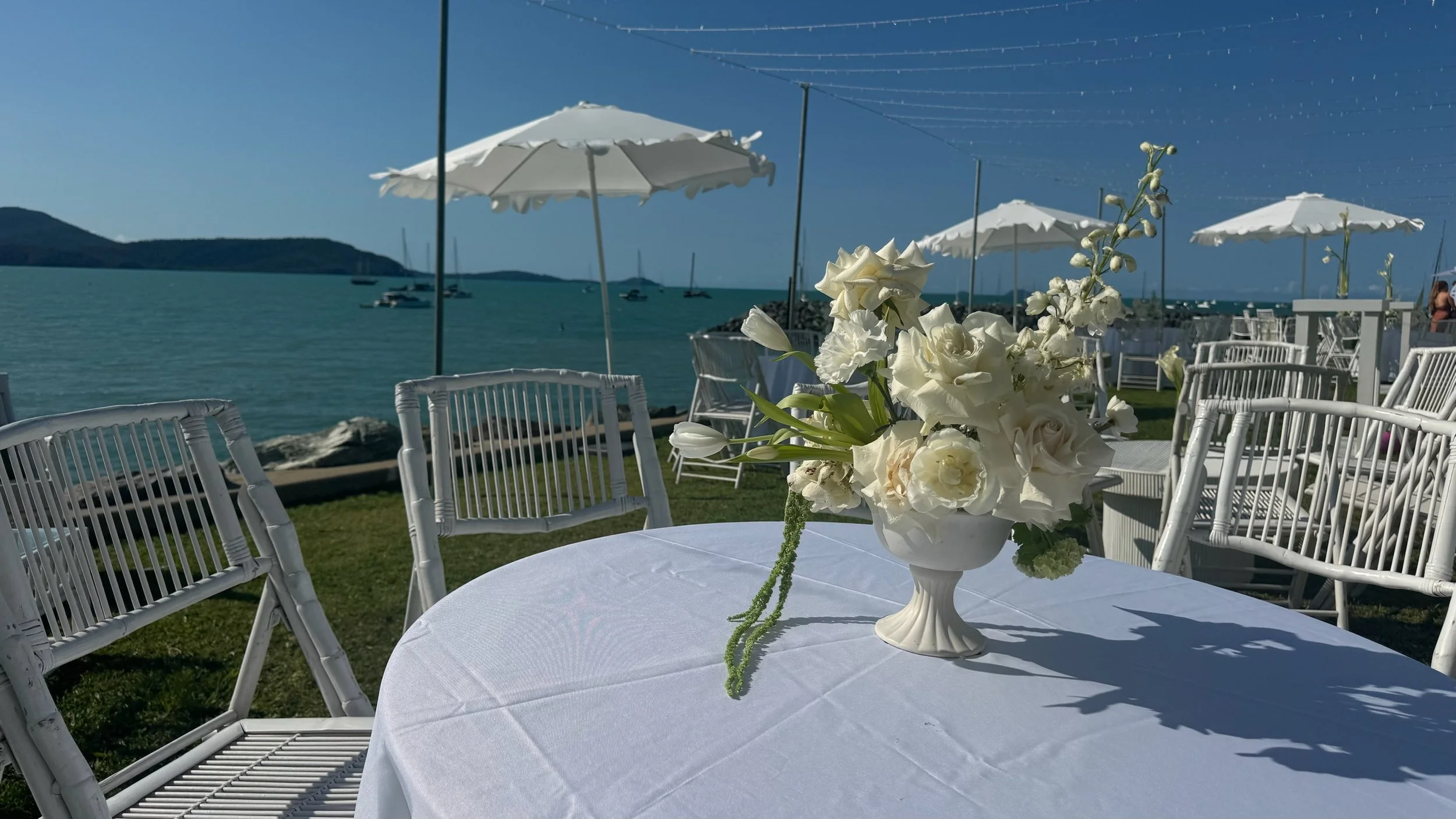 A round table with a white tablecloth and a white flower arrangement in a vase, set outdoors near water with boats in the distance, white umbrellas, and white chairs in the background.