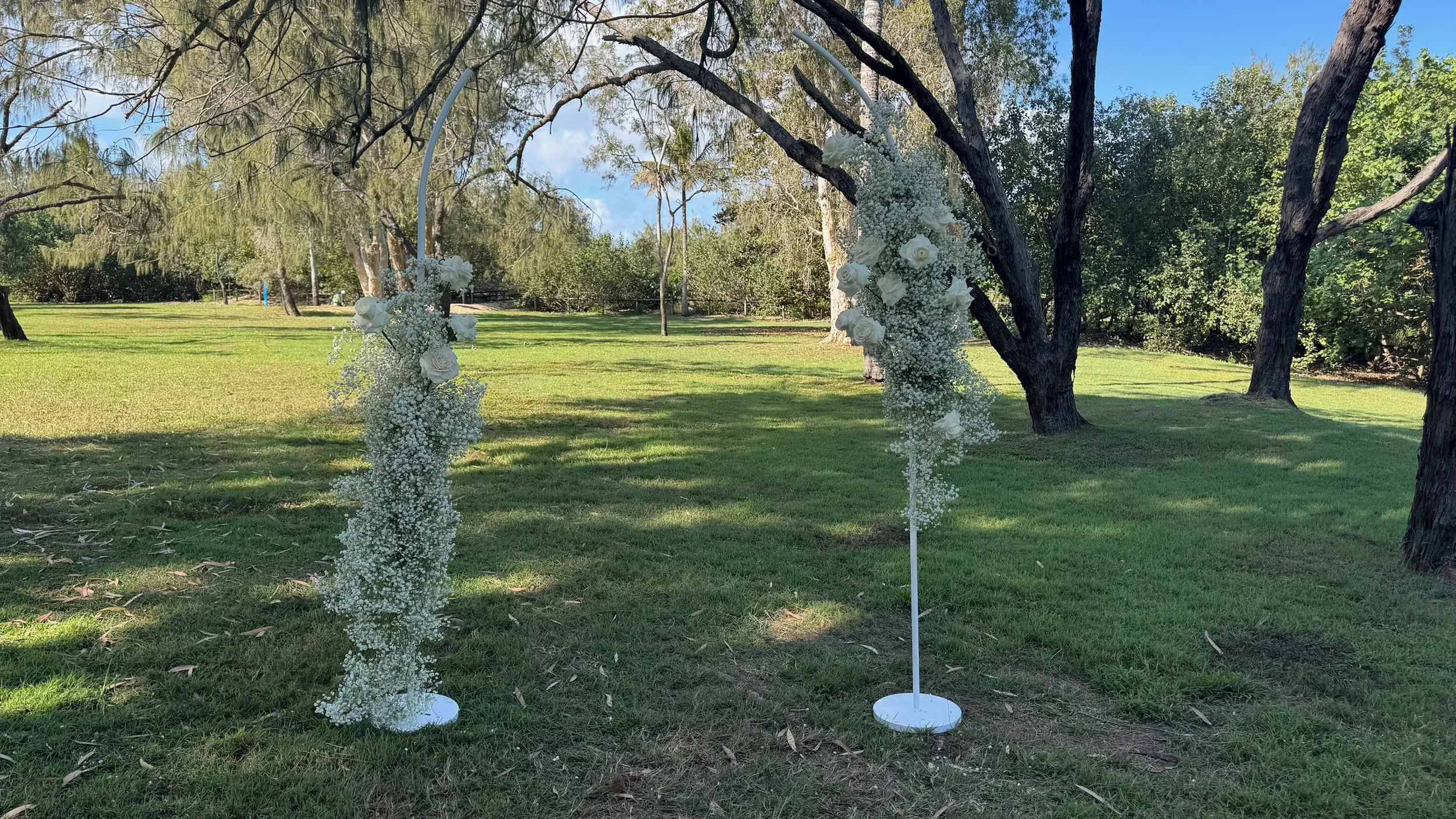 Wedding arch decorated with white flowers and greenery in an outdoor grassy park with trees and blue sky.
