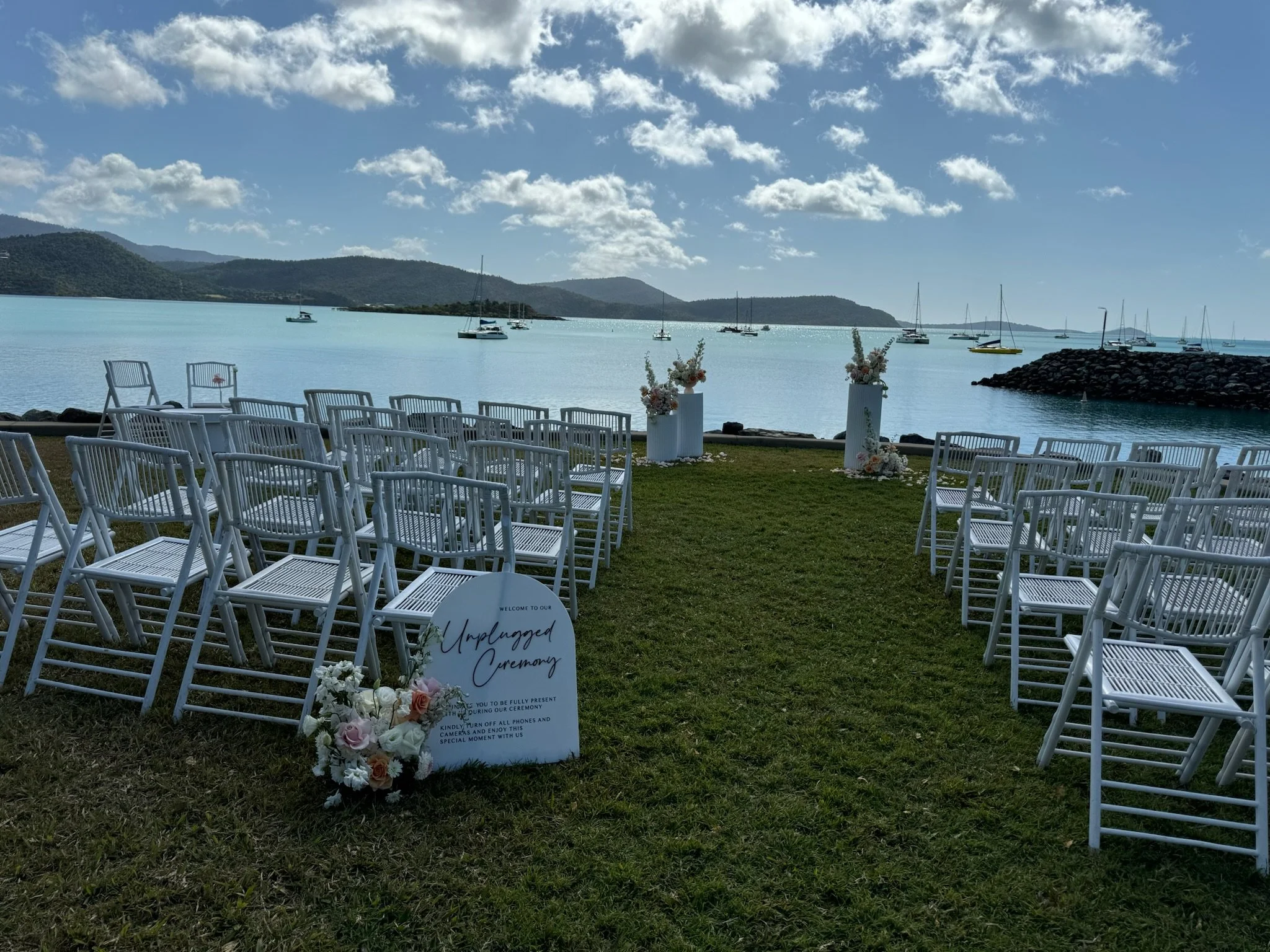 Outdoor wedding ceremony setup with white chairs on grass facing a lake with sailboats, mountains in the background, and a blue sky with scattered clouds. Decor includes flower arrangements and a sign reading 'Unplugged Ceremony'.