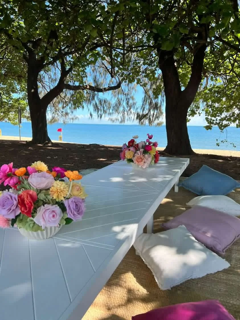 A picnic setup with a white table, several bouquets of colorful flowers, and cushions on a sandy beach under large trees, facing the ocean.