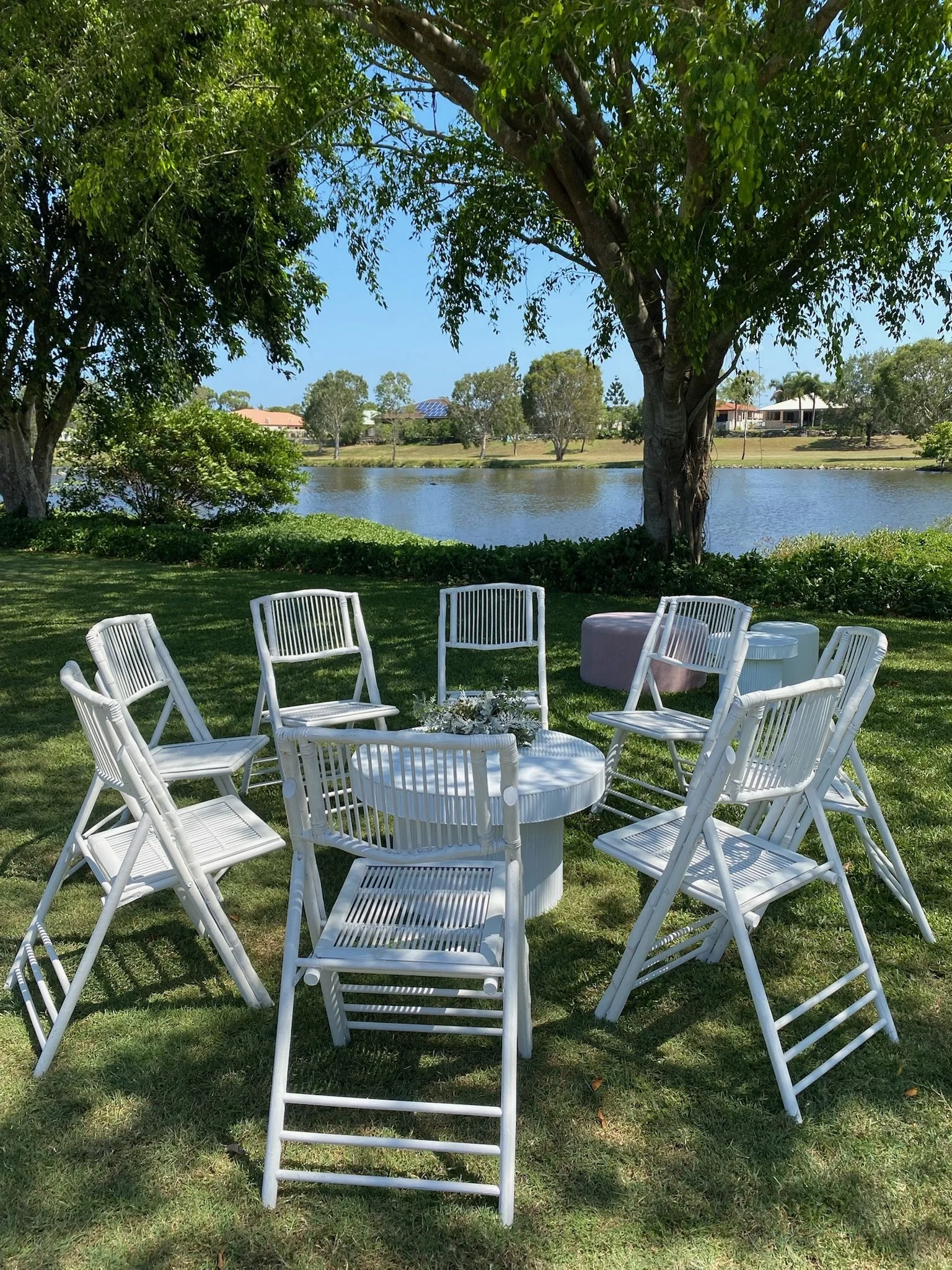 Outdoor seating area with white chairs arranged around a round table by a lake, under a large tree with houses visible in the background.