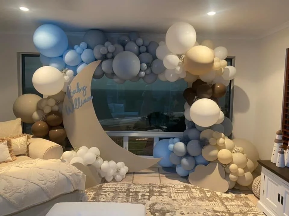Decorative balloon arrangement with a large crescent moon and clouds for a baby shower, featuring blue, white, brown, and beige balloons, and a sign reading "Baby William".