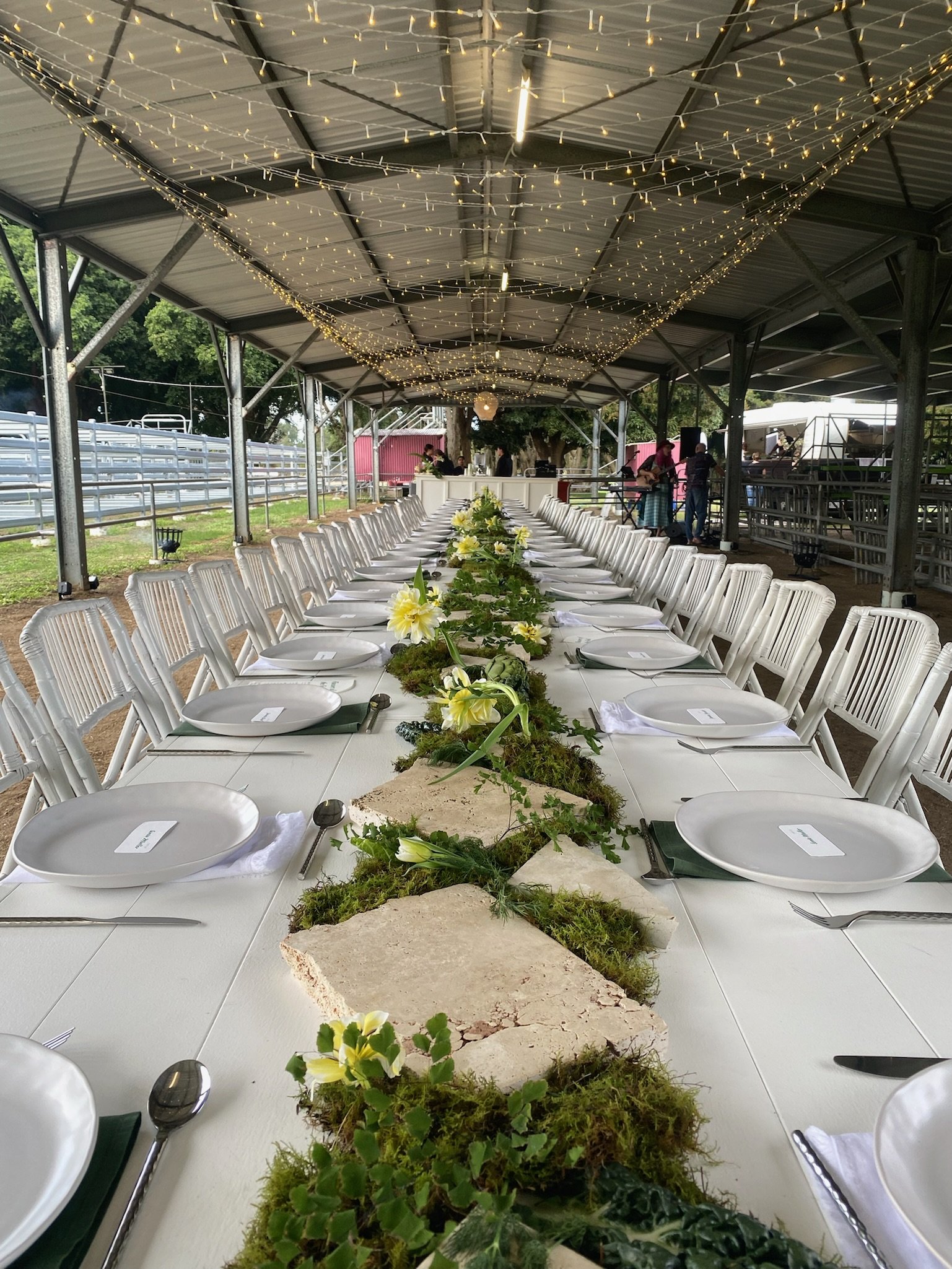 Decorated outdoor banquet table with white plates, silverware, and floral centerpieces under a roof with string lights.