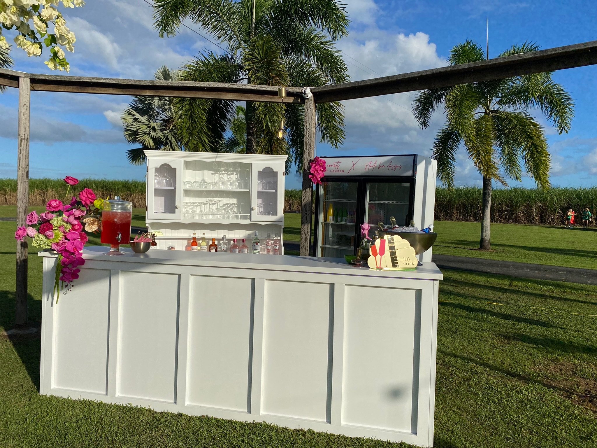 Outdoor white bar cart setup with floral decorations, drink bottles, and a drink dispenser, with palm trees and a grassy field in the background under a partly cloudy sky.