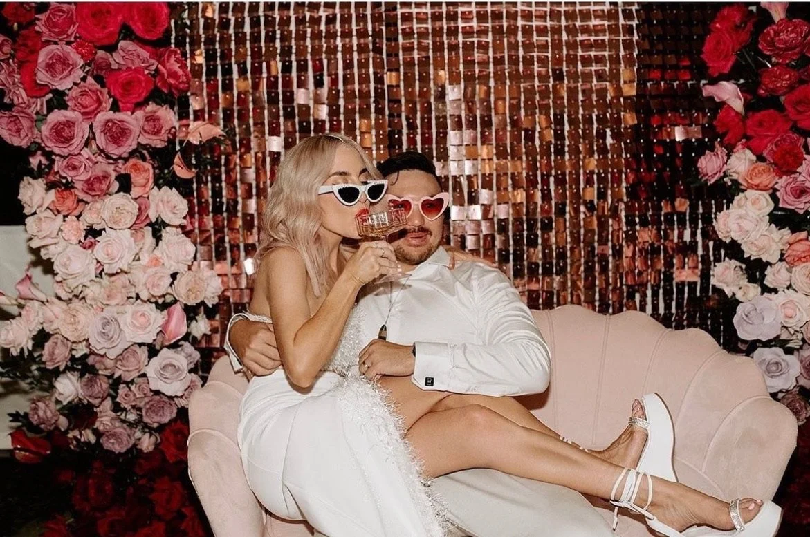 A couple at a wedding or celebration sitting on a pink vintage-style sofa surrounded by pink and red roses, wearing white and beige clothing, with mismatched sunglasses, holding drinks, and enjoying a moment together.