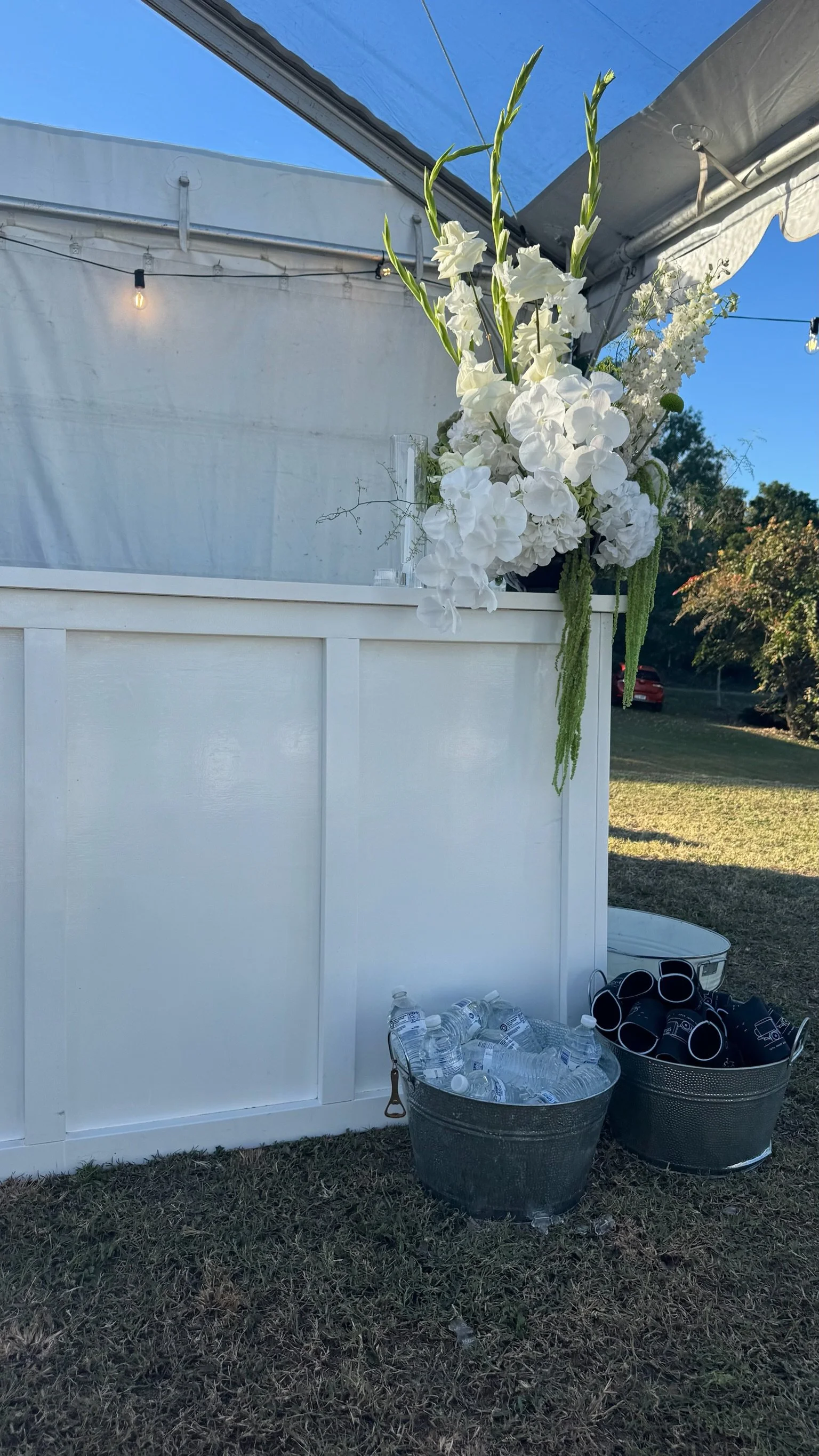 A large floral arrangement with white flowers and greenery on a white bar, two buckets of bottled water and black coffee cups on the ground, outdoor setting with grass and trees under a blue sky.