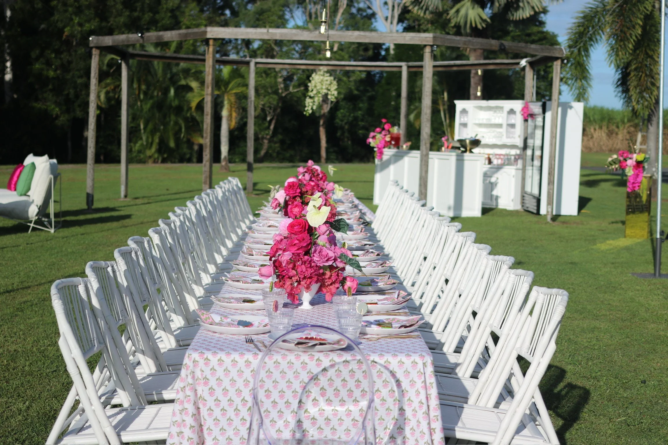 Outdoor wedding reception setup with a long table decorated with pink floral centerpieces, white chairs, and a white bar in the background, on green grass with trees and blue sky.