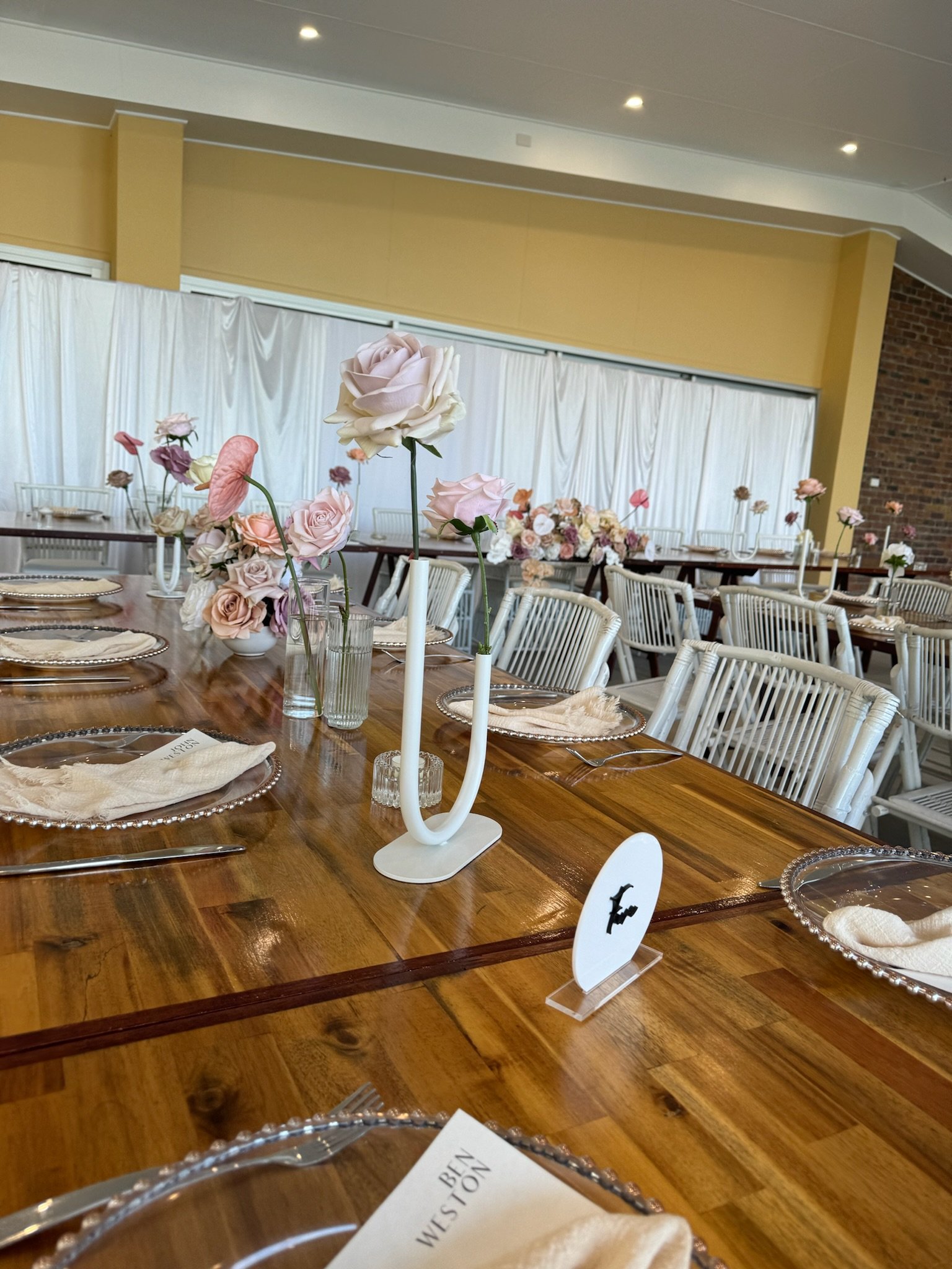 Elegant event table setup with floral centerpieces, candles, silver chargers, and white chairs in a decorated venue.