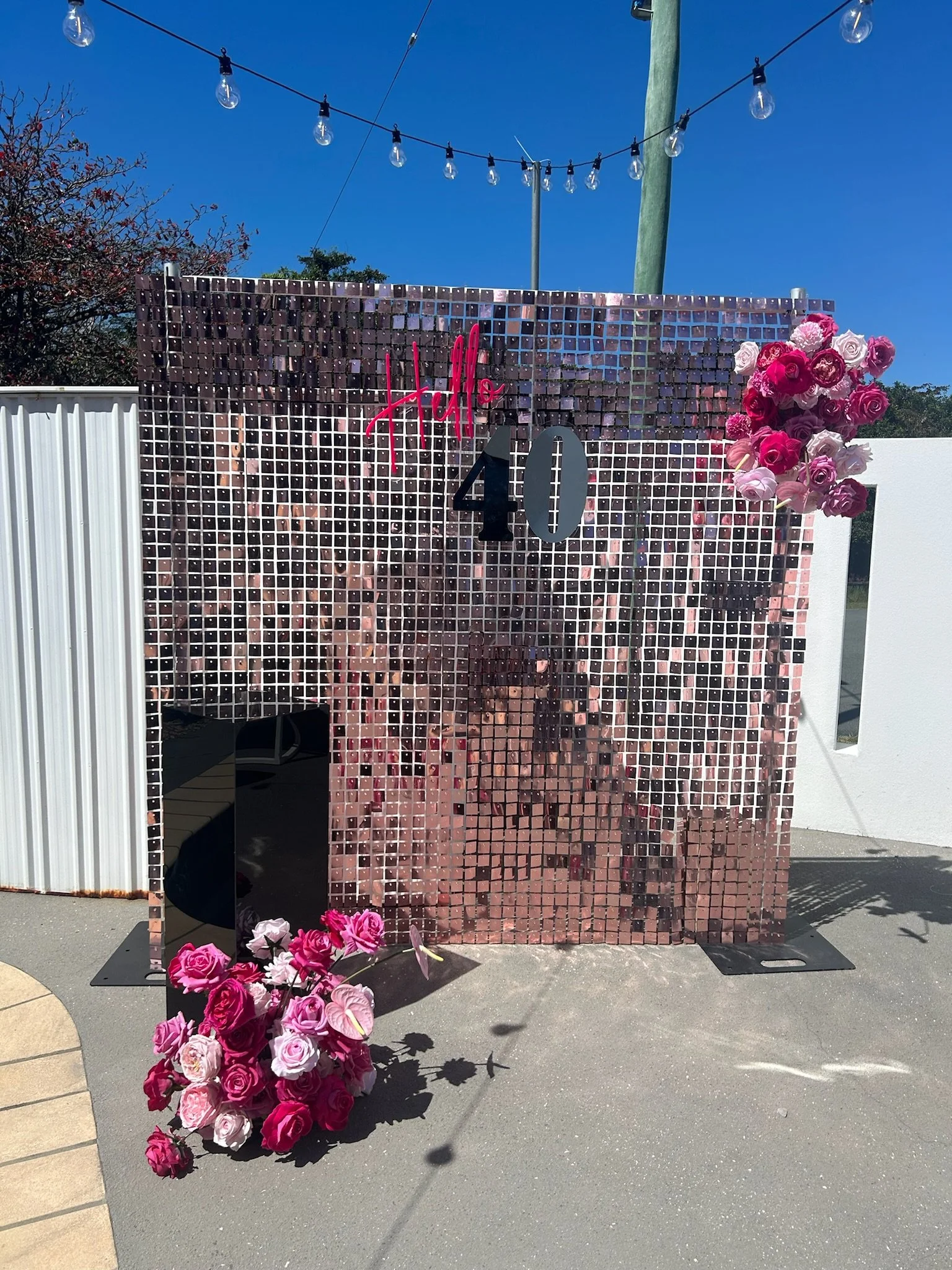 Decorative mirror wall with the words 'Holly 40' and pink and white flowers on the top right and bottom left, with hanging light bulbs above on a sunny day.