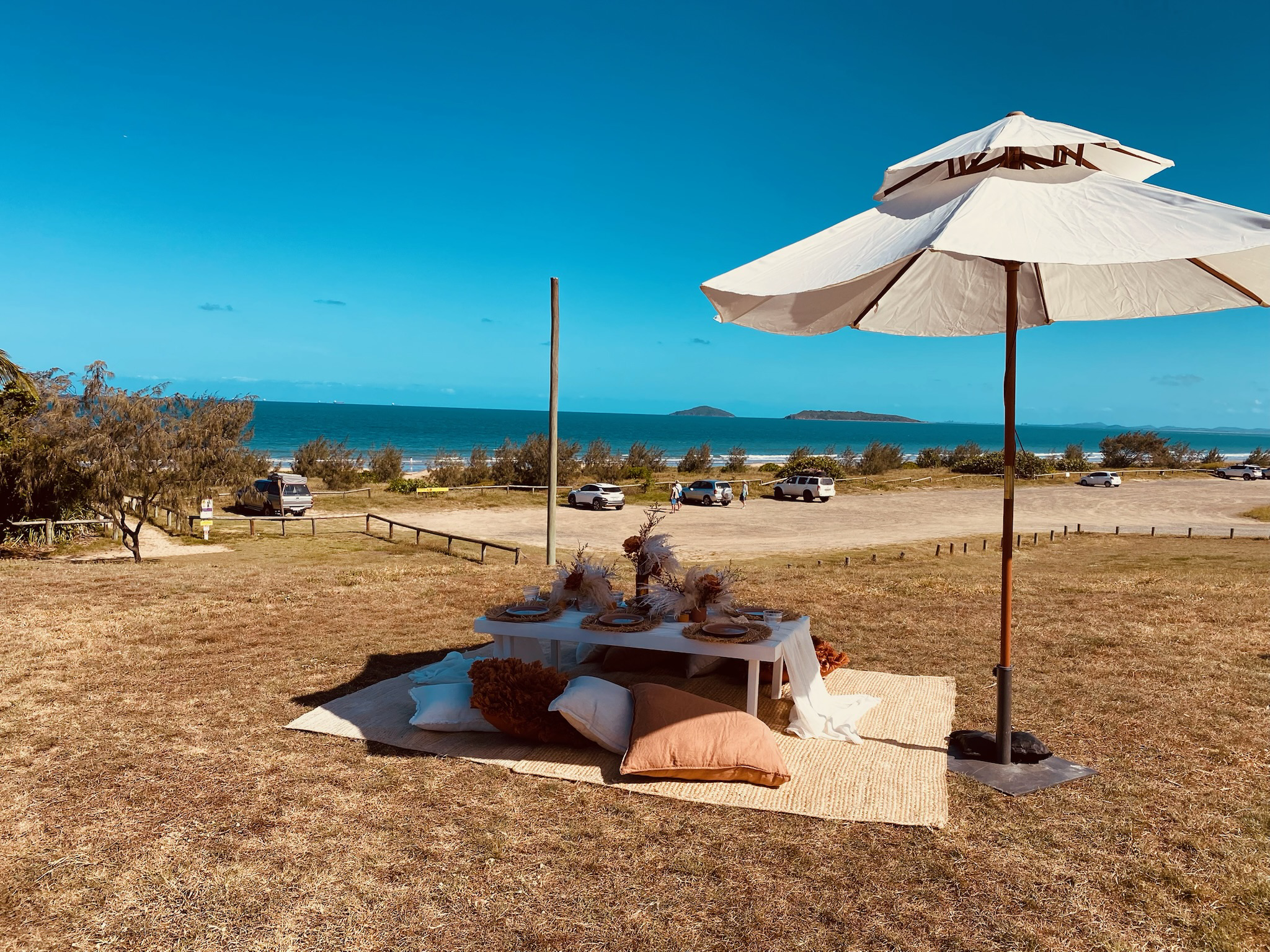 Beachside outdoor setup with a white umbrella, a low table with decorations, pillows, and a rug, overlooking a parking lot, bushes, ocean, and distant islands.
