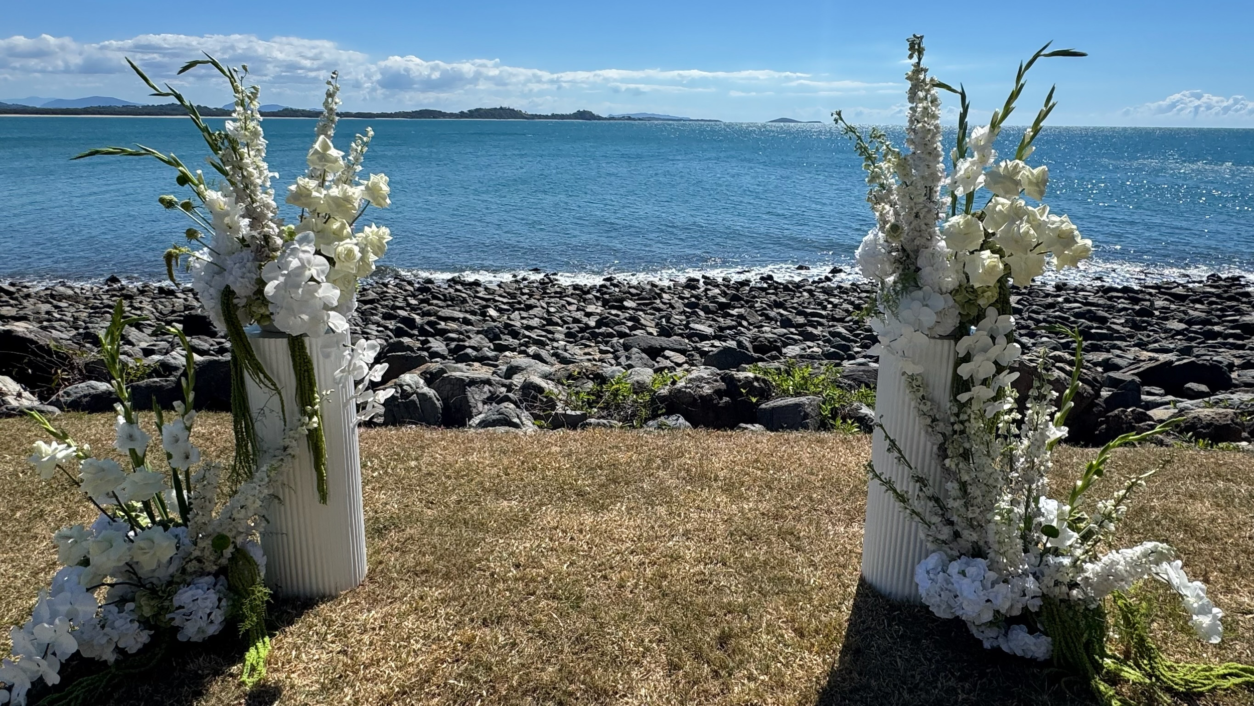 Two white floral arrangements in tall vases on grass by a rocky shoreline, overlooking a blue ocean under a partly cloudy sky.