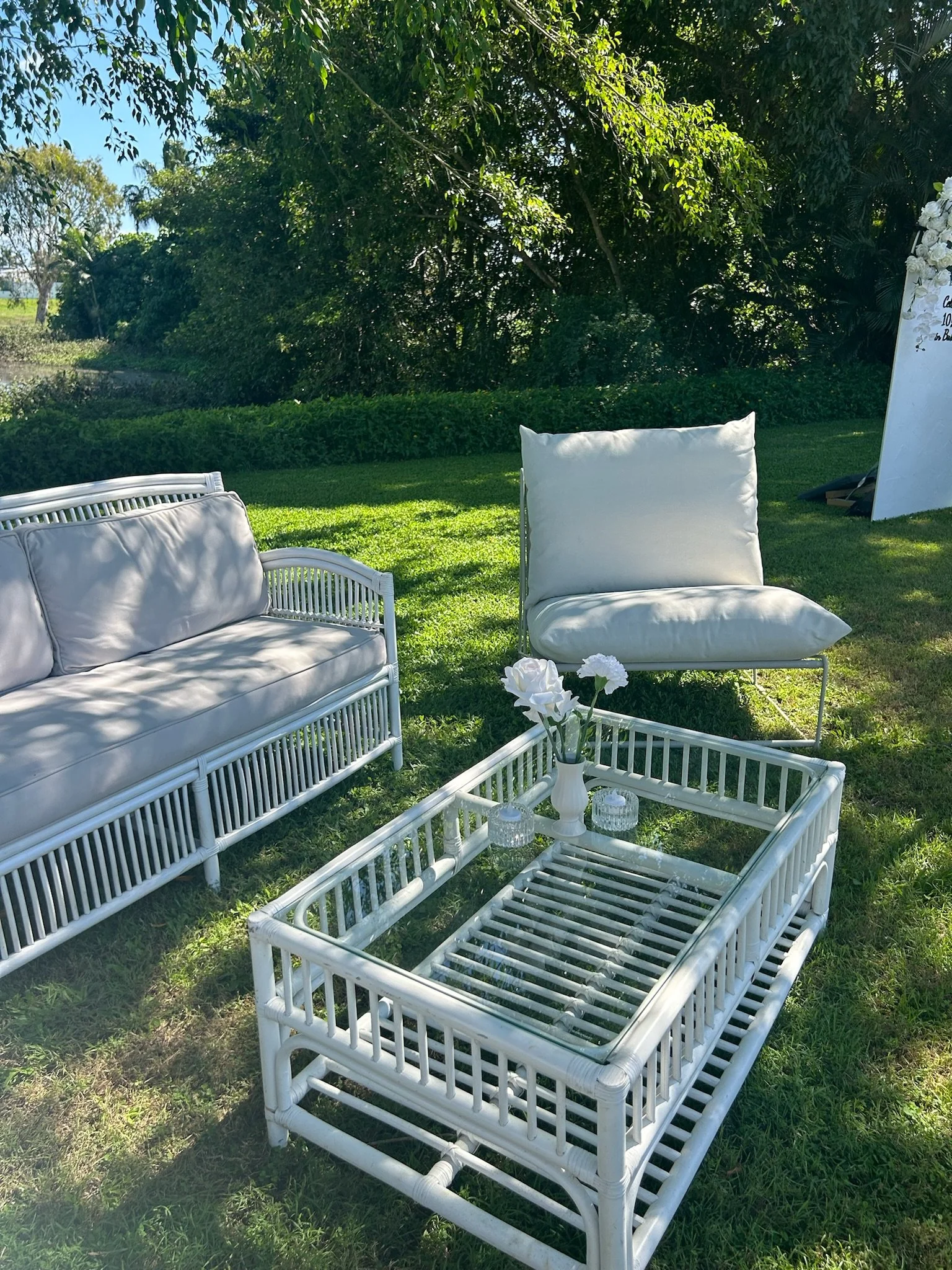 Outdoor seating arrangement with white wicker furniture, including a sofa, a lounge chair, and a coffee table with vases and candles, set on a grassy area with trees and bushes in the background.