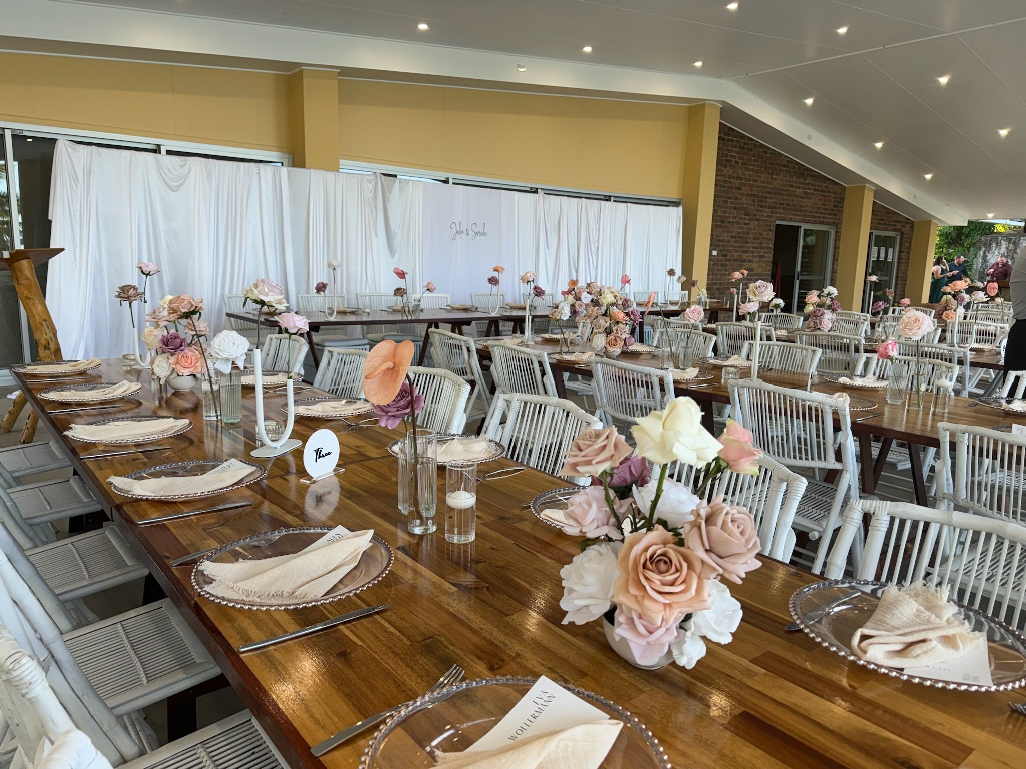 A beautifully decorated indoor event space set for a wedding reception with long wooden tables, white chairs, floral centerpieces in pink, peach, and white tones, and place settings with napkins, plates, and utensils.