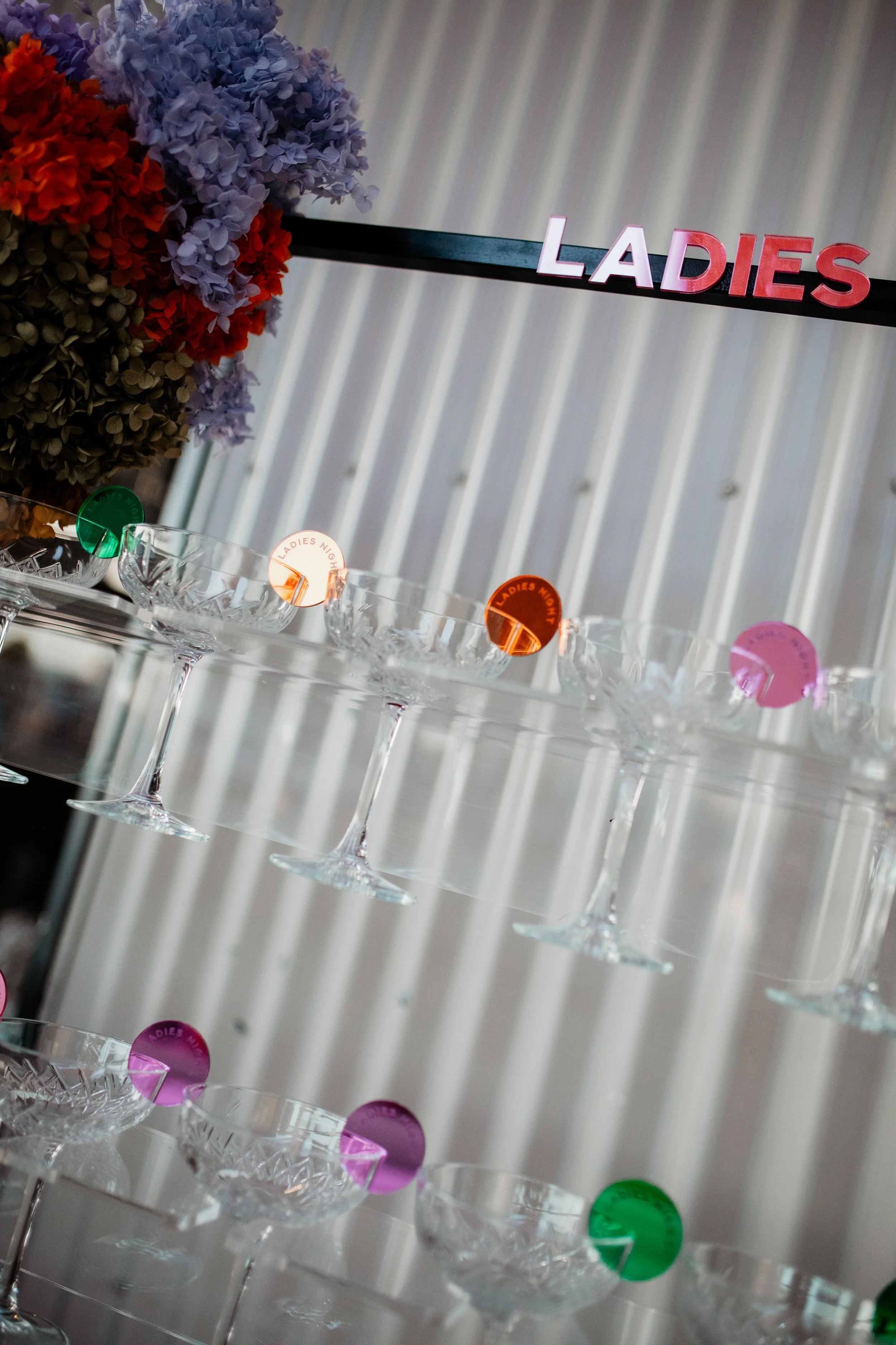 A display of empty coupe glasses with colorful circular stickers, a floral arrangement of purple, red, and brown flowers, and a sign that says 'LADIES'.