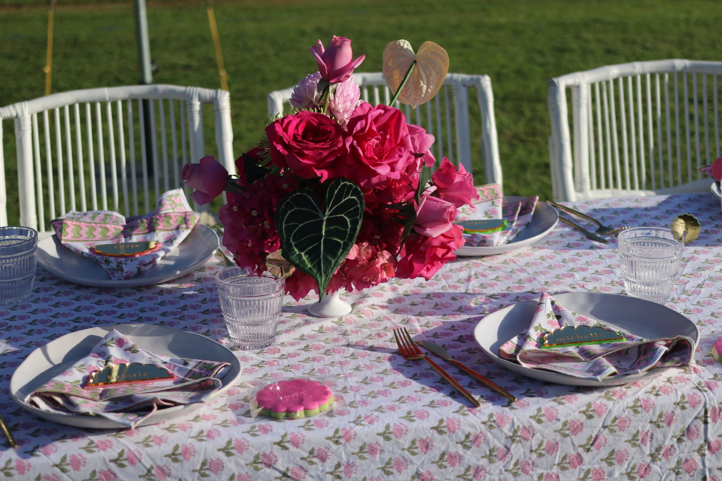 A table set for a gathering with pink and white floral arrangements, white plates with napkins, cutlery, and glasses on a pink floral tablecloth outdoors