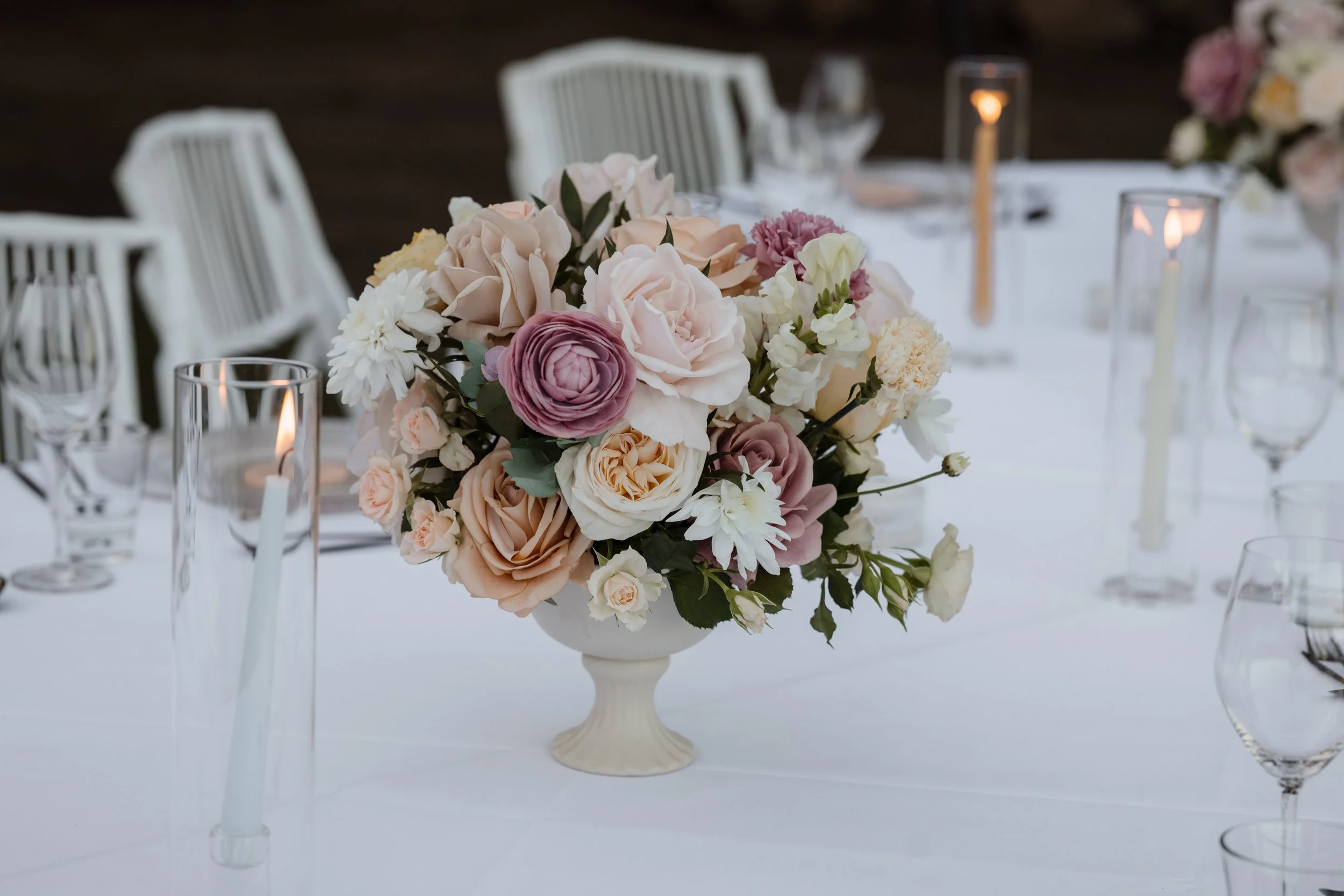 Elegant table centerpiece with pink, white, and blush flowers in a white vase, surrounded by glass candle holders and white chairs, at an outdoor event.