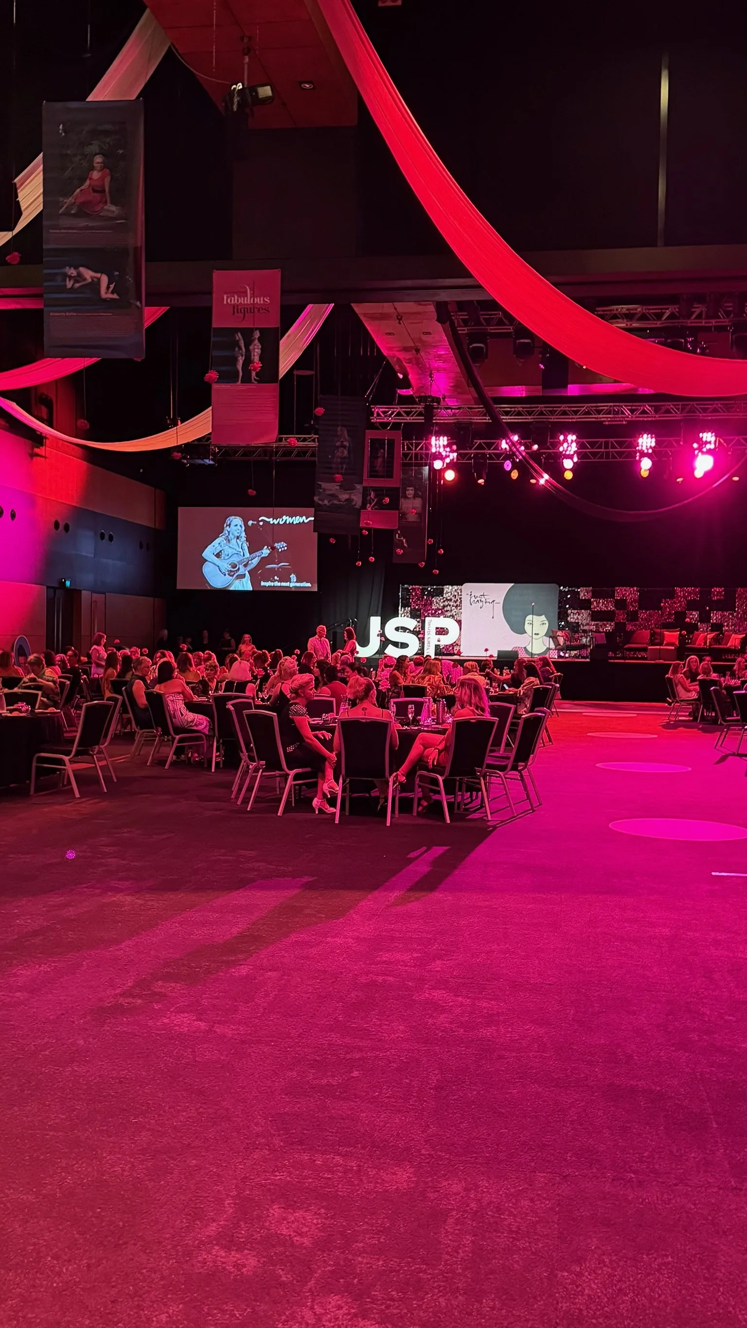 An indoor event space with pink lighting, round tables with seated guests, a stage with large letters "JSP," and a woman playing guitar projected on a screen.