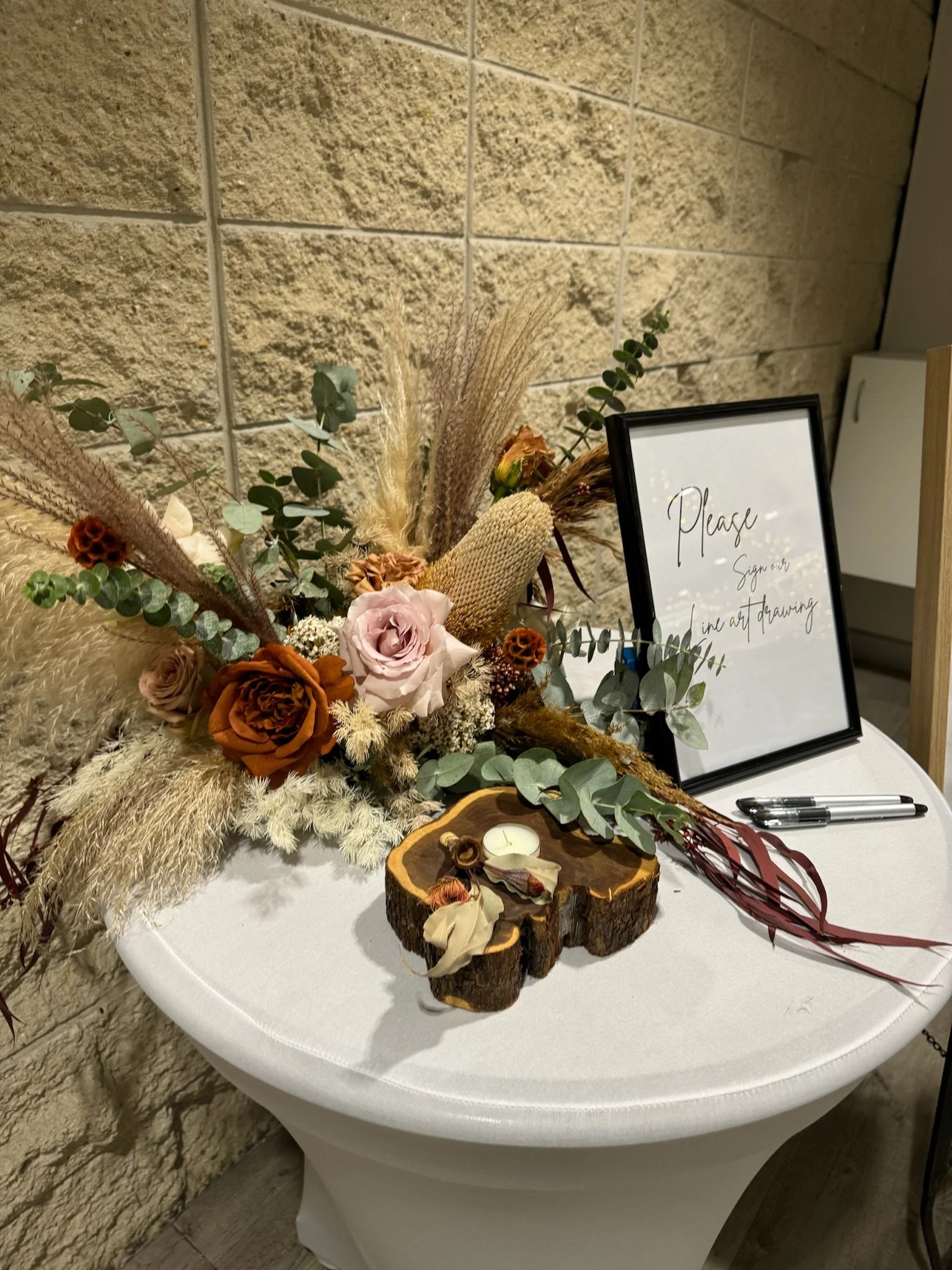A white round table with a flower arrangement of dried and fresh flowers, a framed sign that says "Please Sign or in the guest art drawing," a black marker, a small candle in a wood slice holder, and some decorative ribbons against a brick wall backg