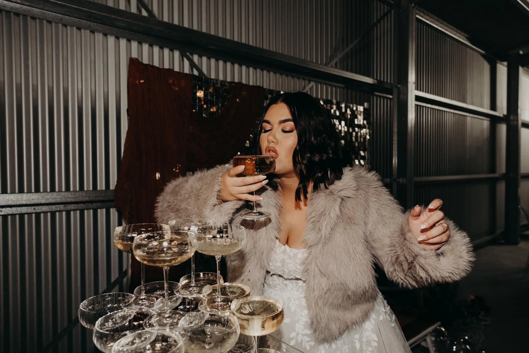 A woman with dark hair in a beige fur coat and a white dress drinks champagne from a coupe glass in a metallic industrial-style setting, with several other champagne glasses in front of her.