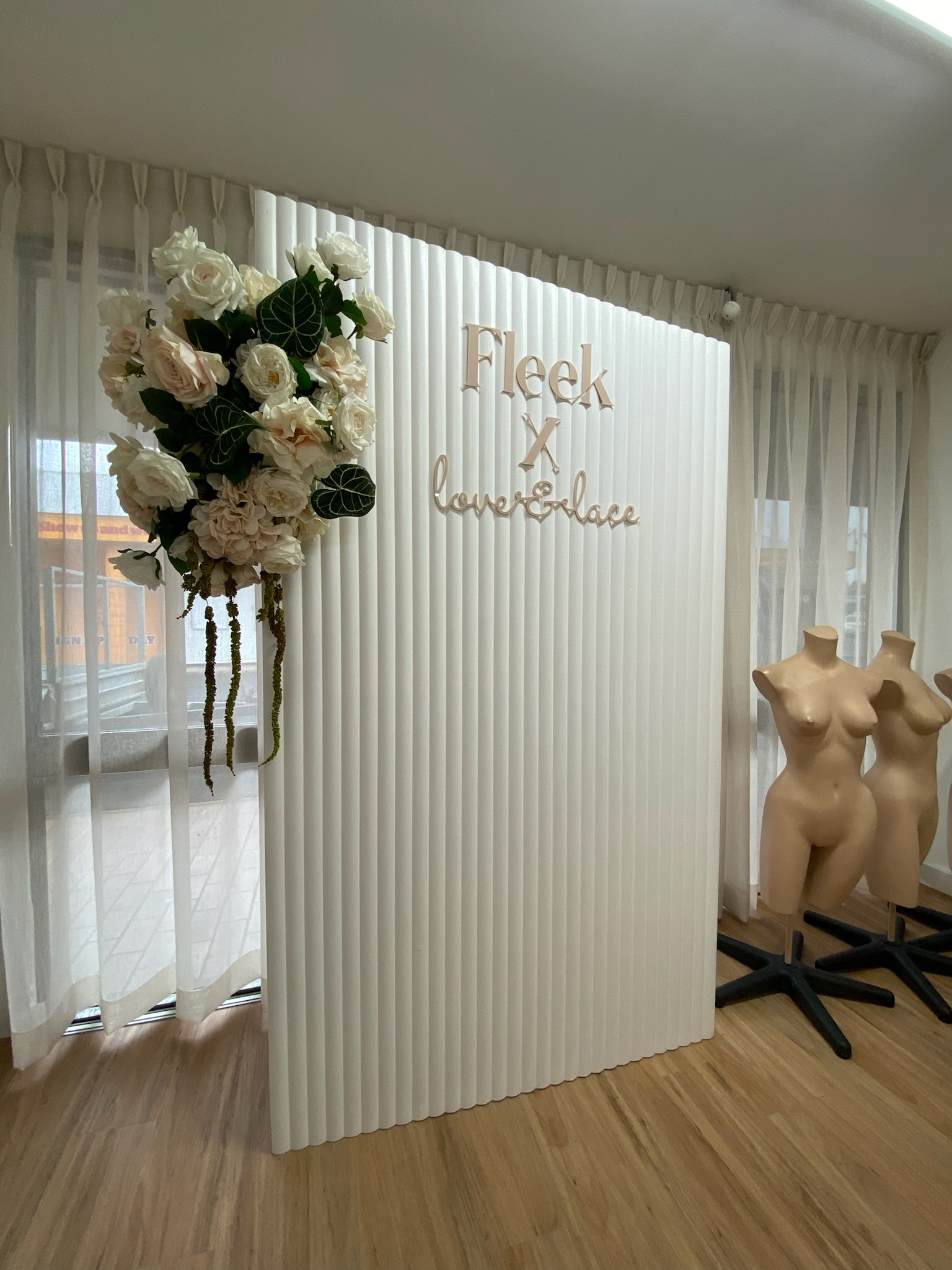 White decorative wall with floral arrangement on the left and a sign that reads 'Fleek x love & lace,' with two mannequin torsos on pedestals to the right.