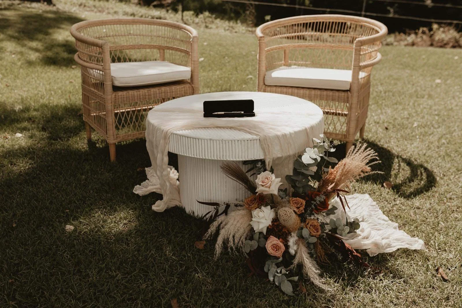 Two wicker chairs with white cushions are positioned on either side of a round table with a white tablecloth in an outdoor setting. The table has a black case or box on top, and a flower arrangement with roses, eucalyptus, and dried grasses is placed