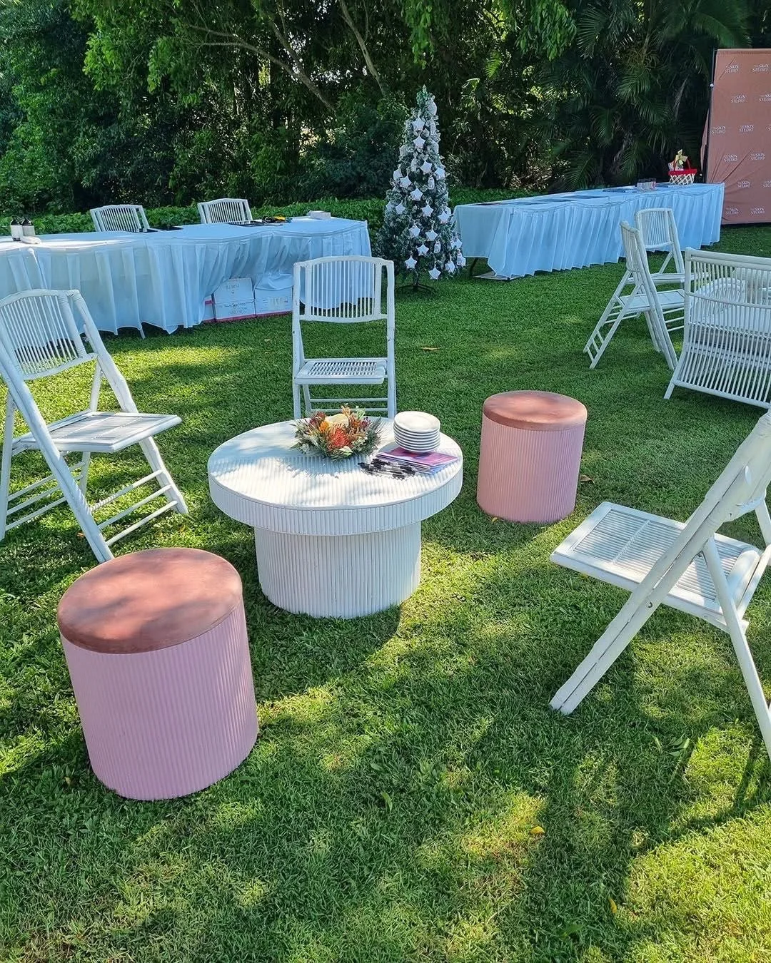 Outdoor event setup with white chairs, pink stools, a round white table with a floral arrangement, and long tables covered with white tablecloths, on a grassy lawn with trees and decorations in the background.